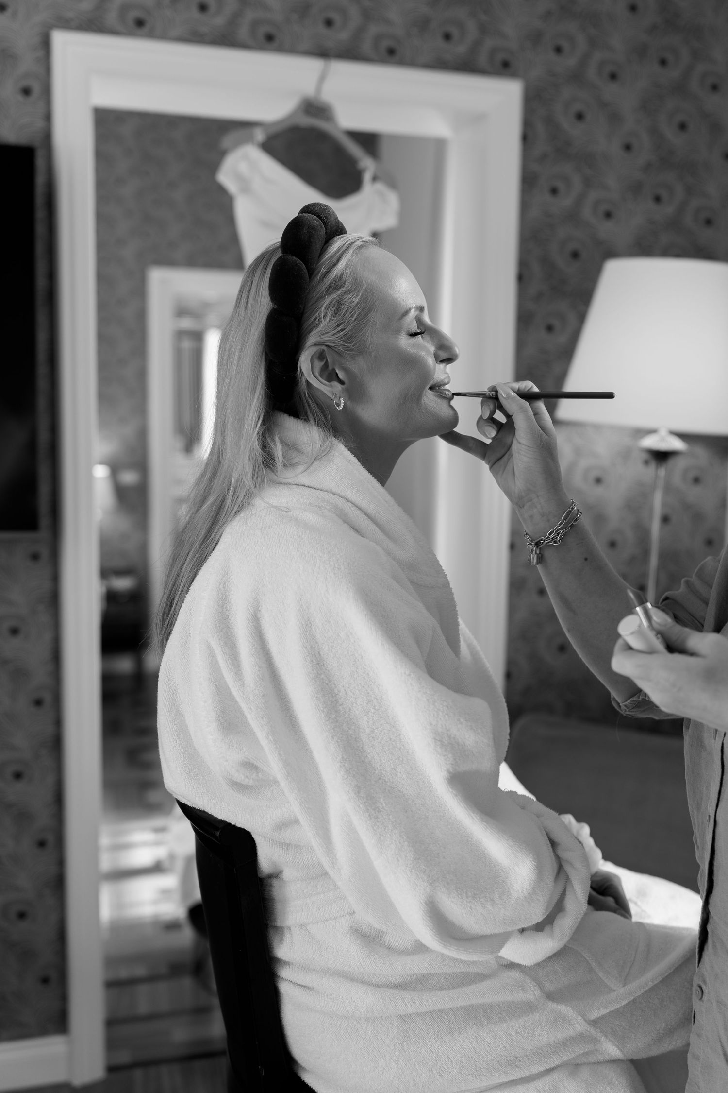 Black and white photo of a woman in a robe getting makeup applied by a professional artist before a wedding.