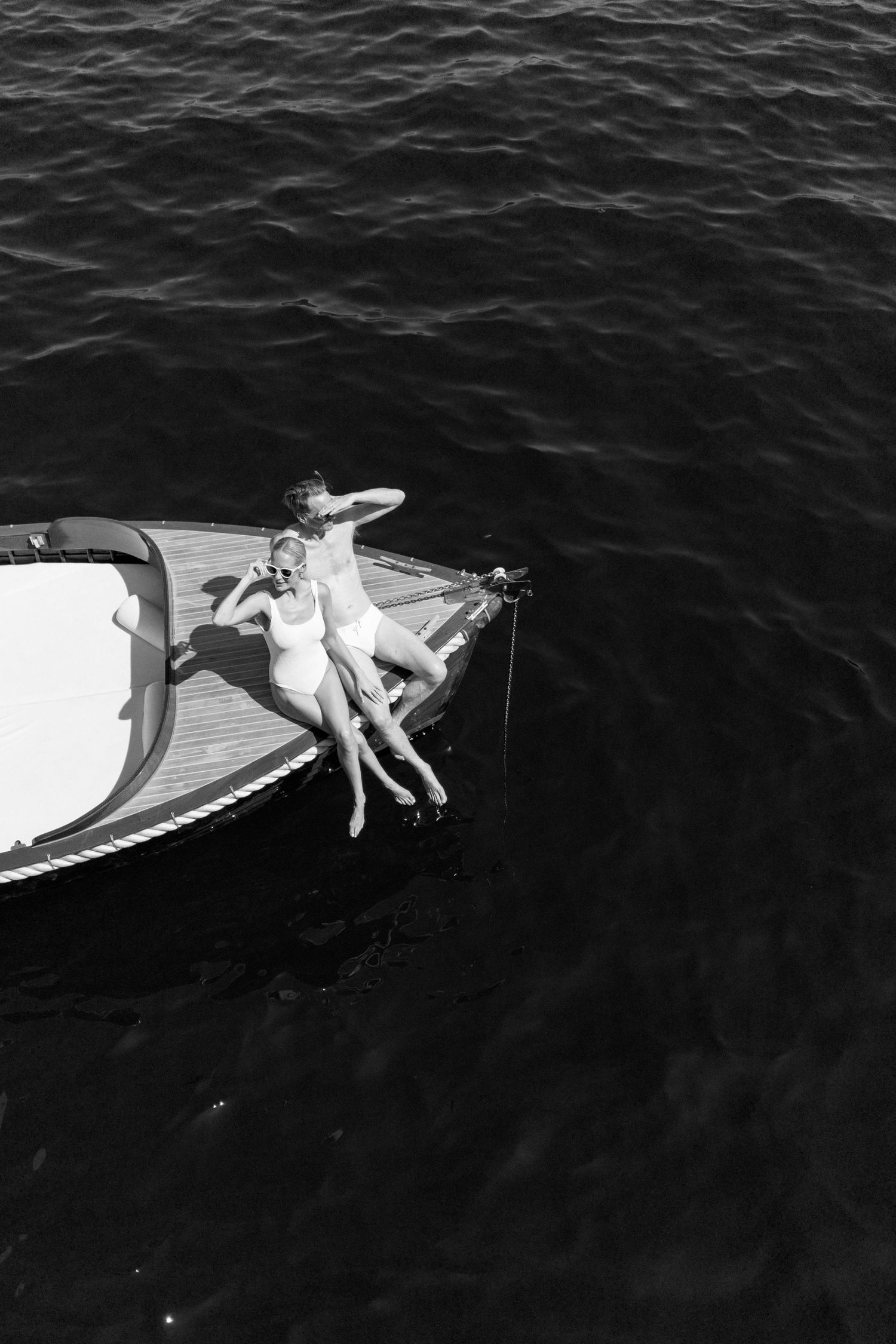 Aerial black and white photo of a couple sunbathing on the bow of a boat surrounded by dark water.