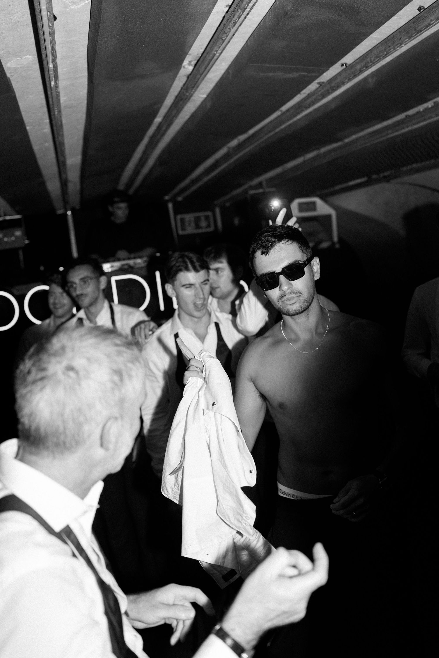 Shirtless man in sunglasses dances with guests in a dark crowded wedding reception venue.