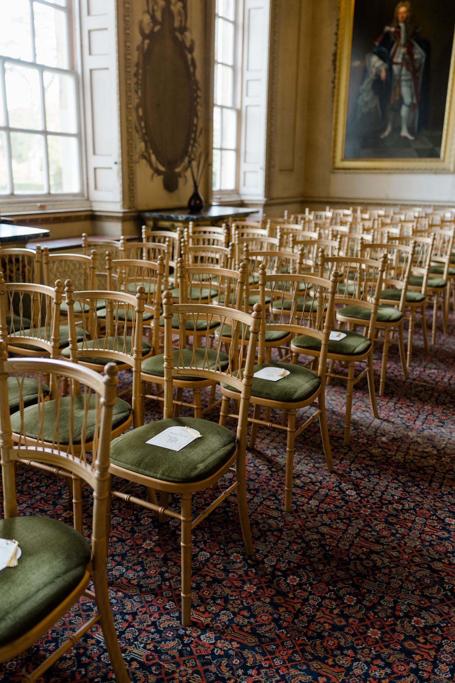 Elegant gold chiavari chairs with green velvet cushions arranged in rows on ornate carpet in a grand historic venue.