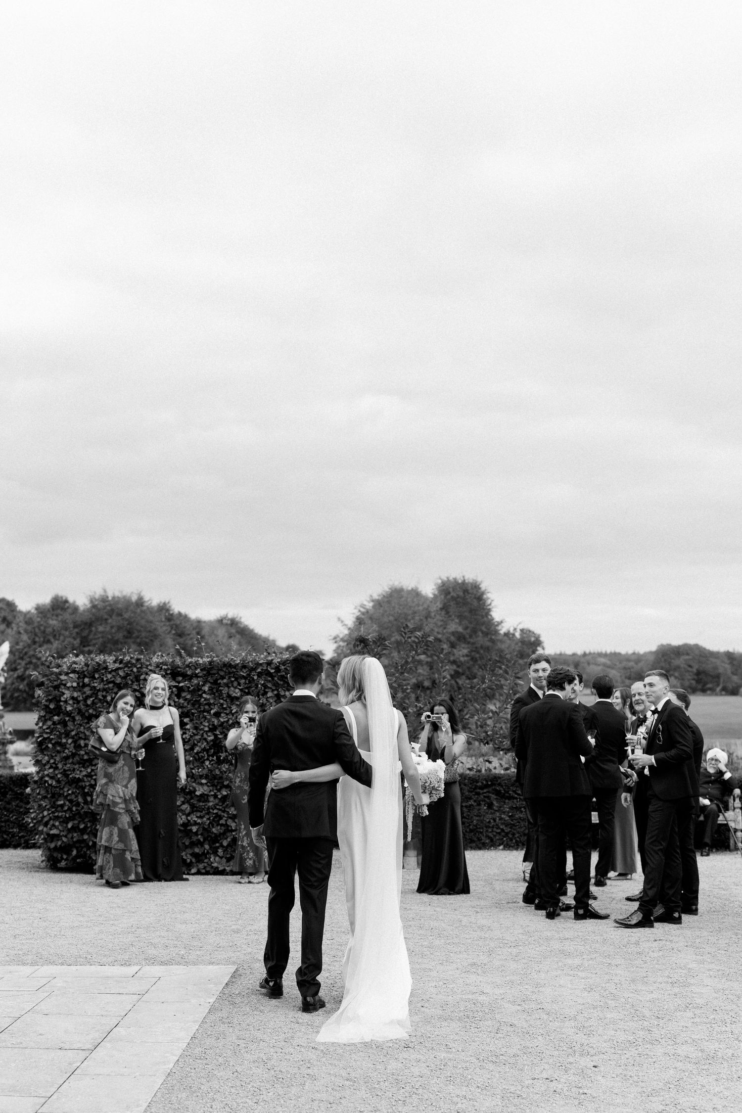 Black and white photo of bride and groom standing together at outdoor wedding ceremony with guests mingling in background.