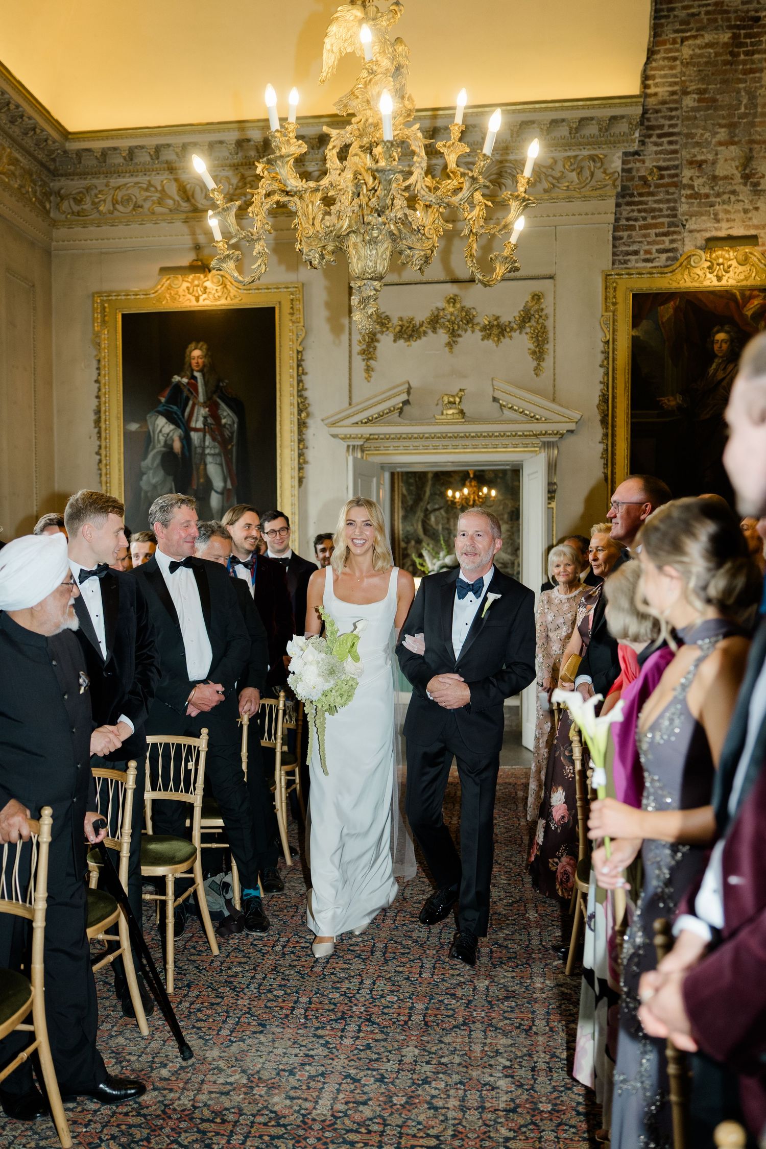 Bride walks down the aisle in elegant white gown at a grand venue with chandelier and oil paintings.