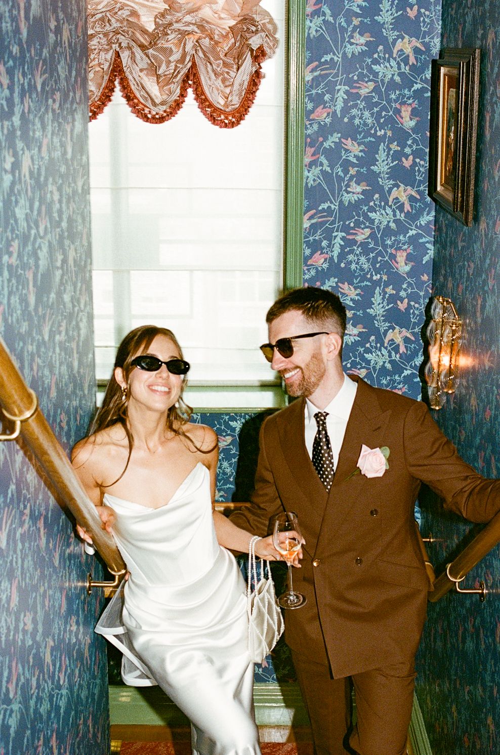 Stylish newlyweds wearing sunglasses laugh together on decorated staircase at wedding reception.