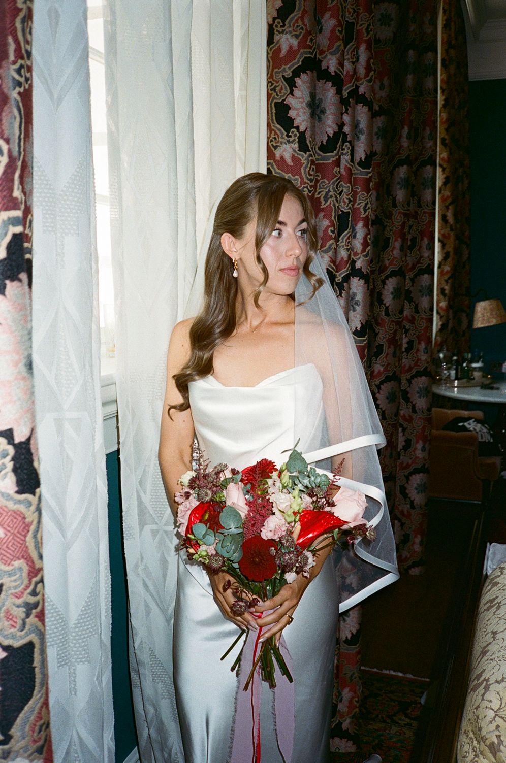 Bride in strapless white gown holding red and pink bouquet with veil, standing by window with floral curtains.