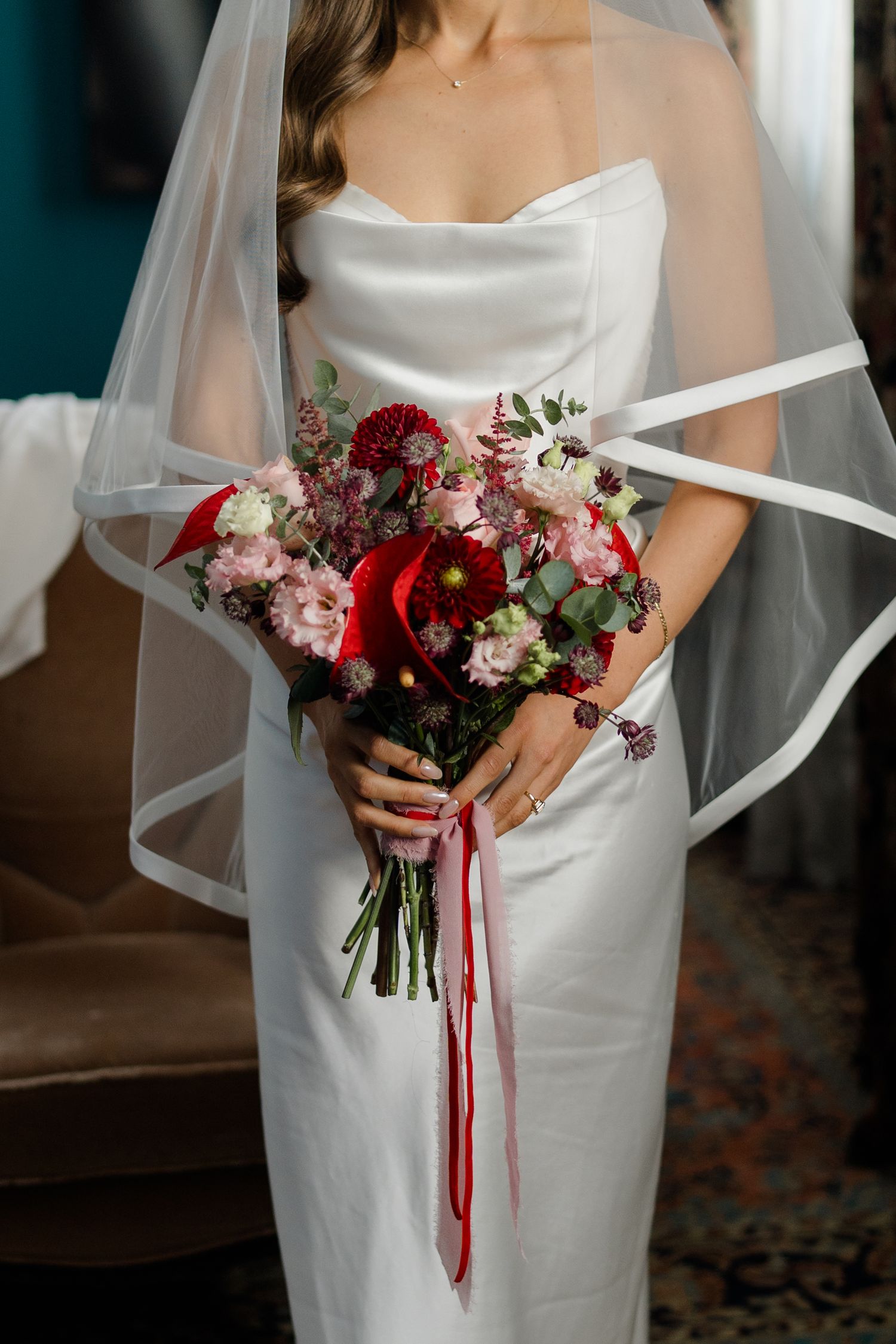 Bride in white strapless gown holding red and pink wedding bouquet with ribbon streamers and veil draped over shoulder.