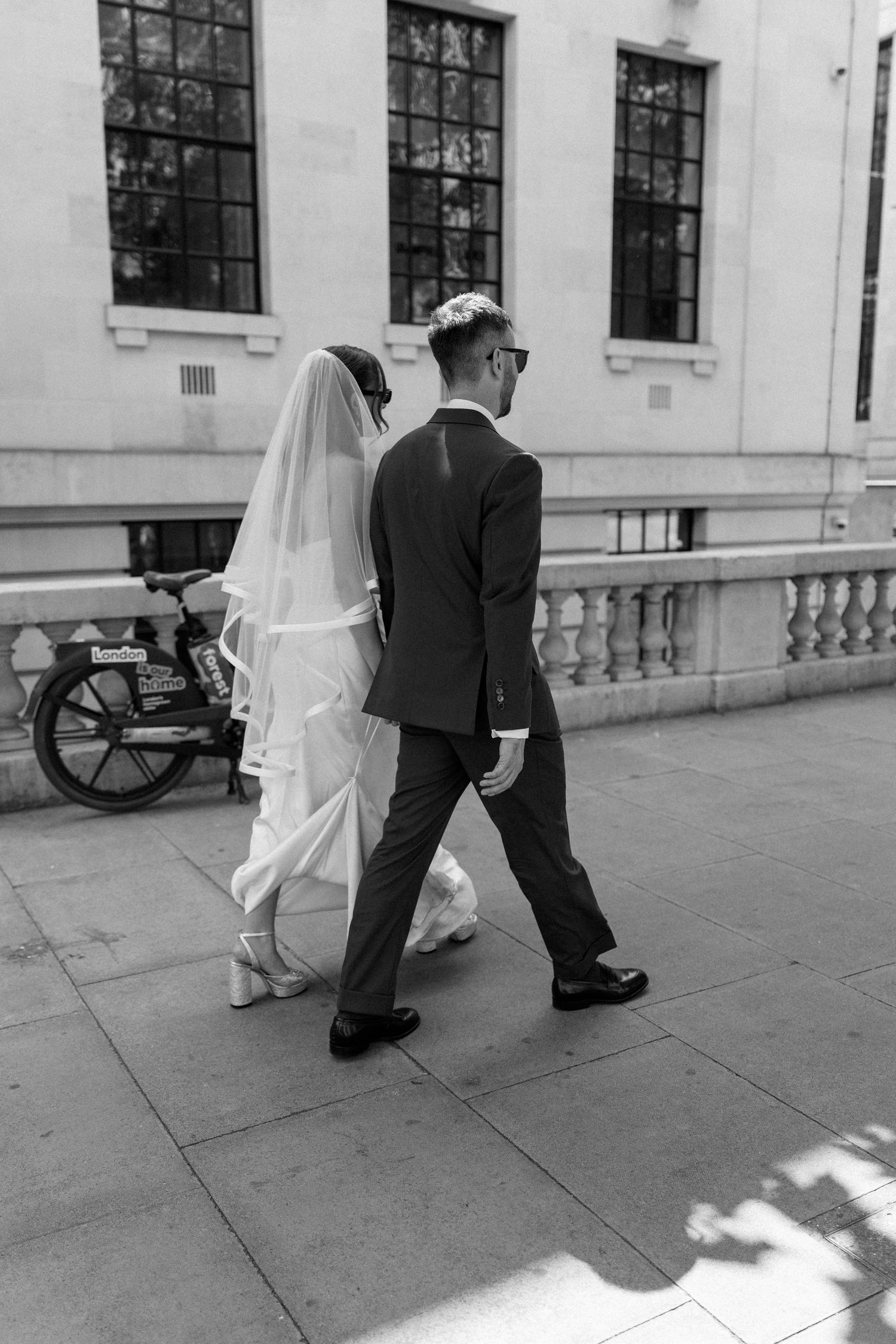 Black and white photo of bride and groom walking past a classical building on their wedding day.