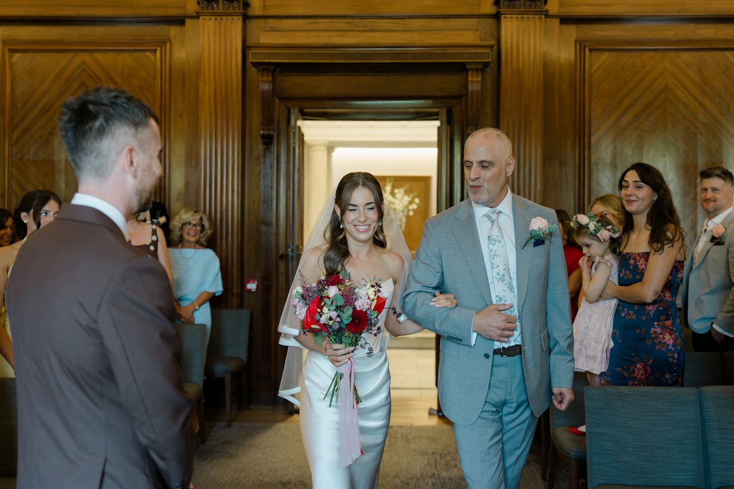 Bride walks down the aisle with her father, holding a red bouquet, as groom waits at a elegant wedding venue.