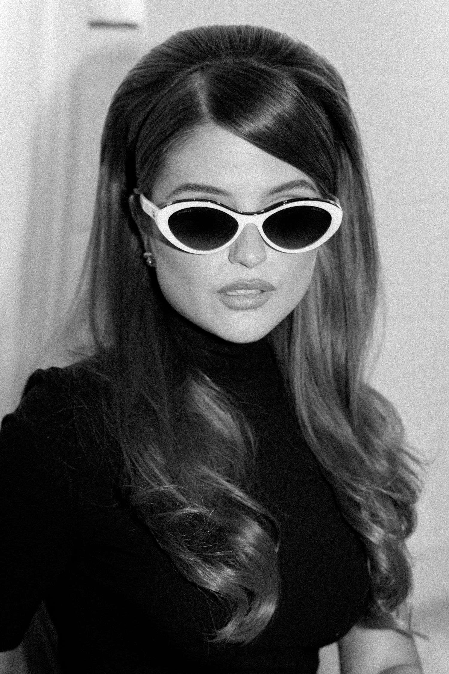 Vintage black and white photo of a young woman with voluminous hair wearing cat-eye sunglasses and black turtleneck.