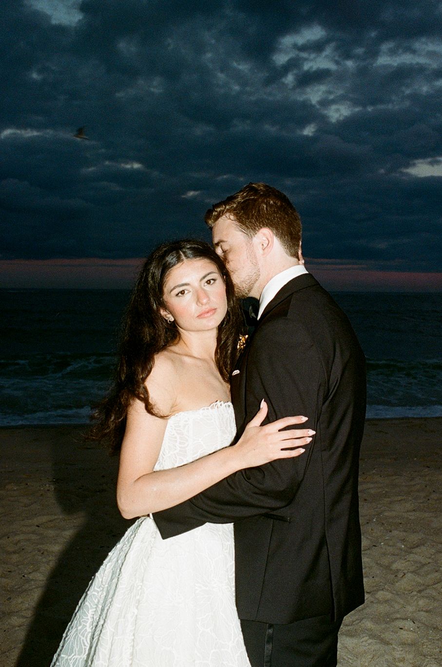 Bride and groom embrace on a beach at dusk with dramatic dark clouds, bride in white strapless gown, groom in black tuxedo.