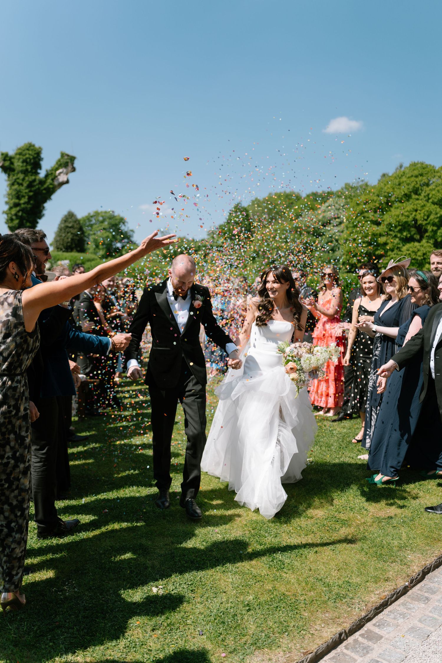 Wedding celebration in a lush garden with guests throwing confetti into the bright blue sky.