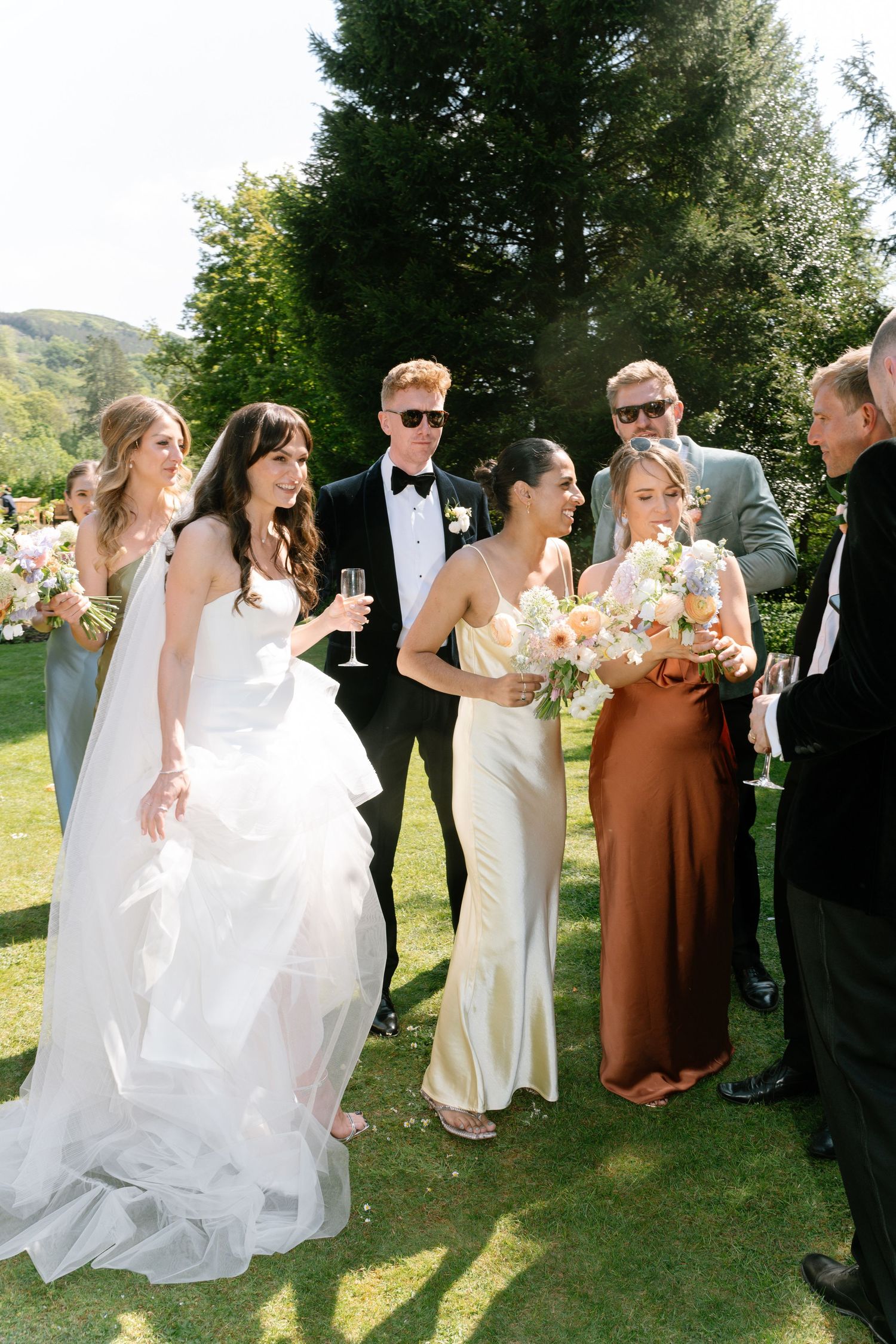 Wedding party in formal attire gathers on a lawn with mountain scenery and floral bouquets.