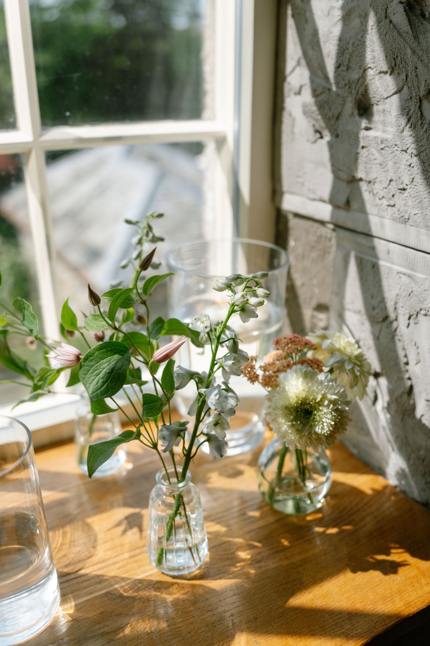 Fresh flowers and greenery in glass vases sit on a wooden table by a sunlit window.