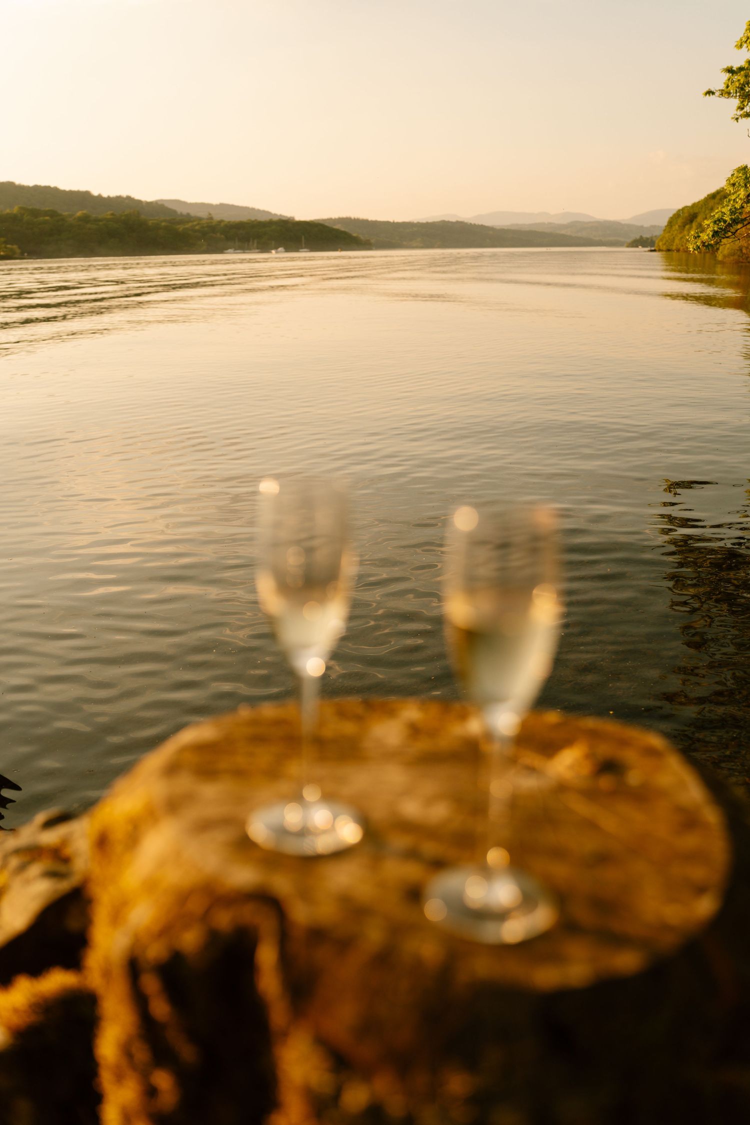 Two champagne glasses sit on a rock overlooking a serene lake at sunset.