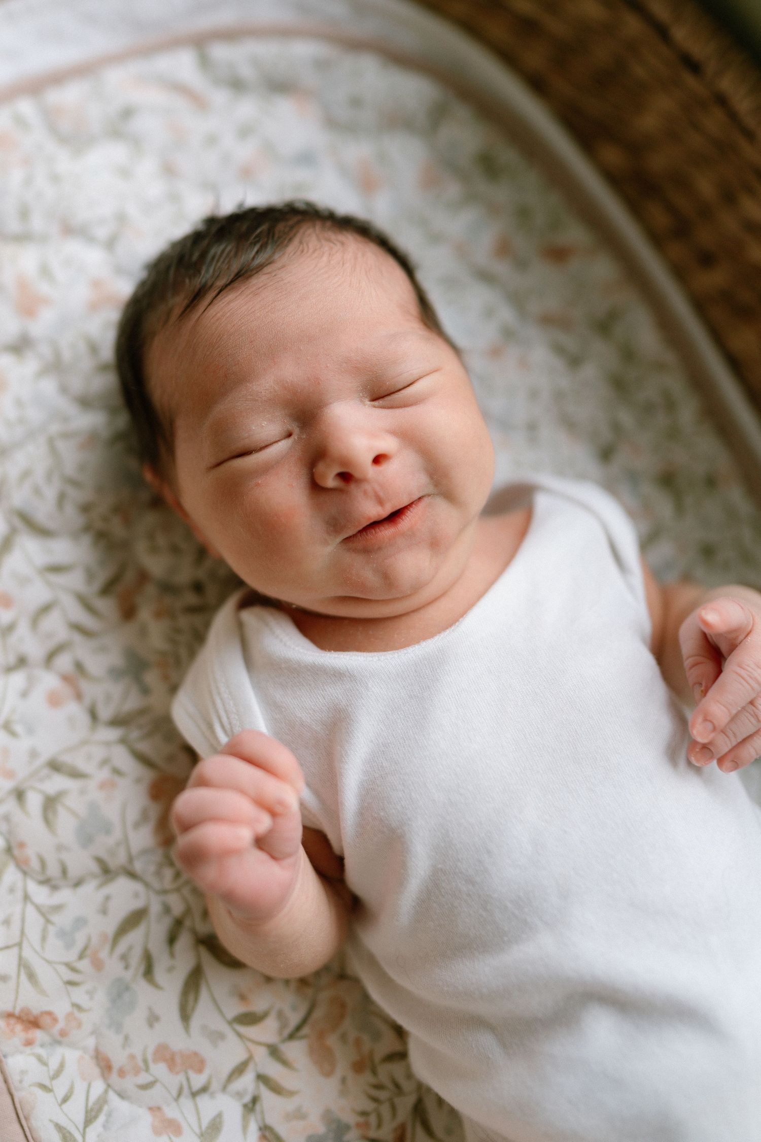 Peaceful newborn baby wearing white sleeper smiling while sleeping on floral fabric.