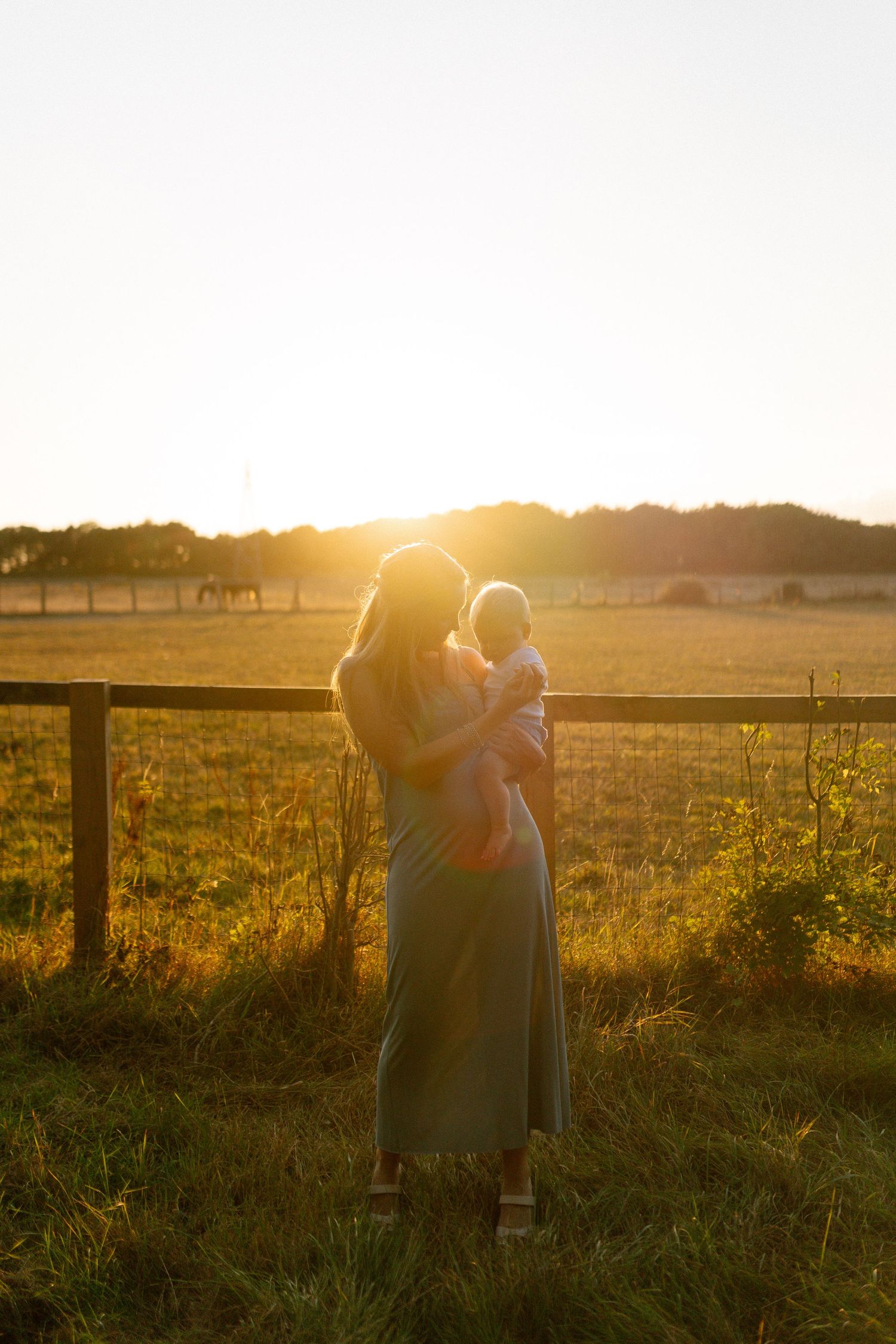 Silhouettes embrace against a golden sunset in a pastoral field with wooden fencing.