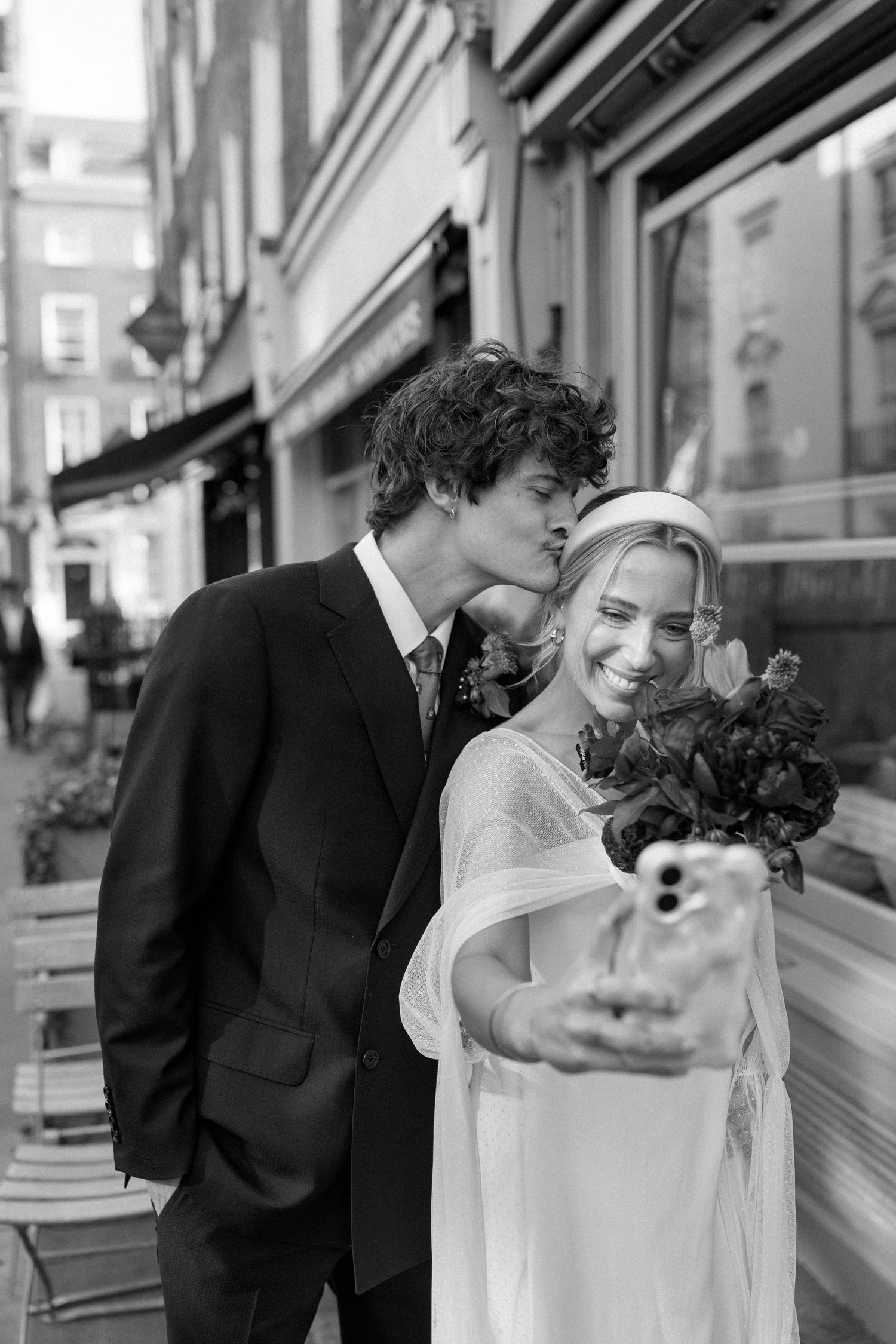 Black and white photo of newlyweds taking selfies together on a city sidewalk.