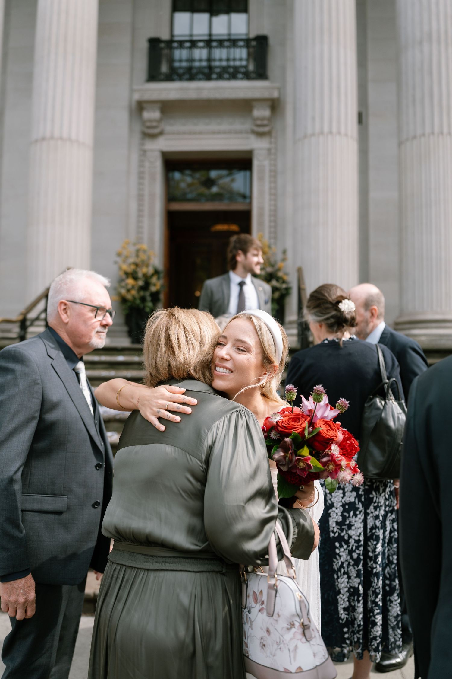 Wedding guests embracing outside classical building with floral bouquet.