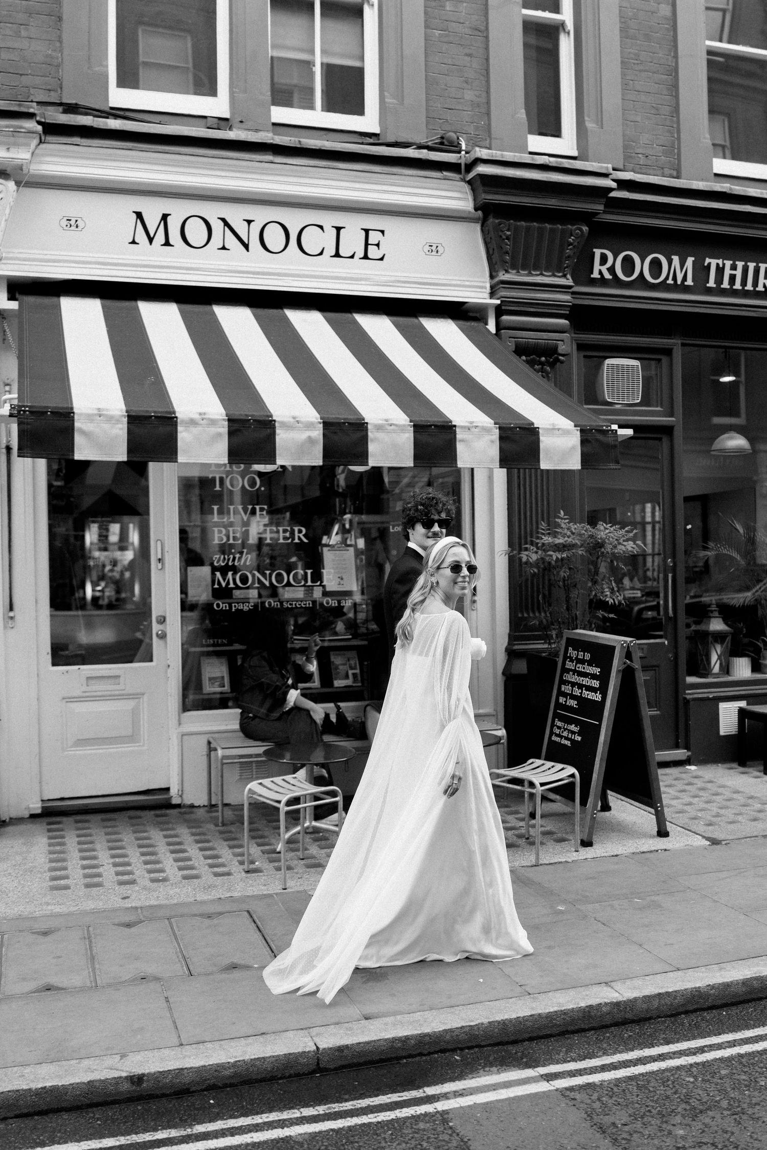 A bride in flowing white dress walks past Monocle store with striped awning.