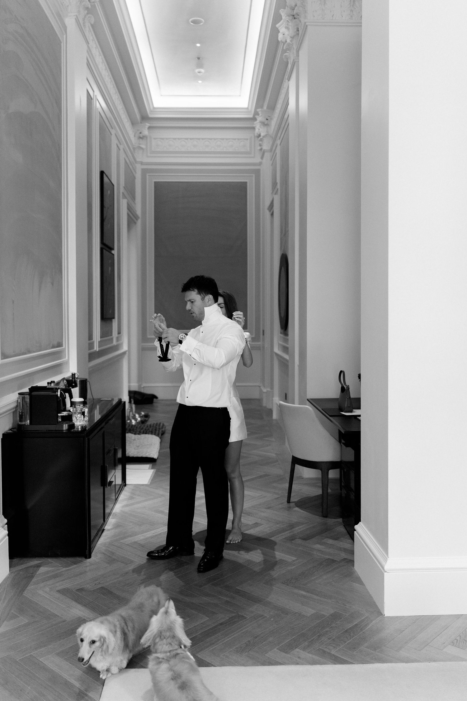 Black and white photo of a man adjusting his cufflinks in an elegant hallway with a dog resting on the floor.