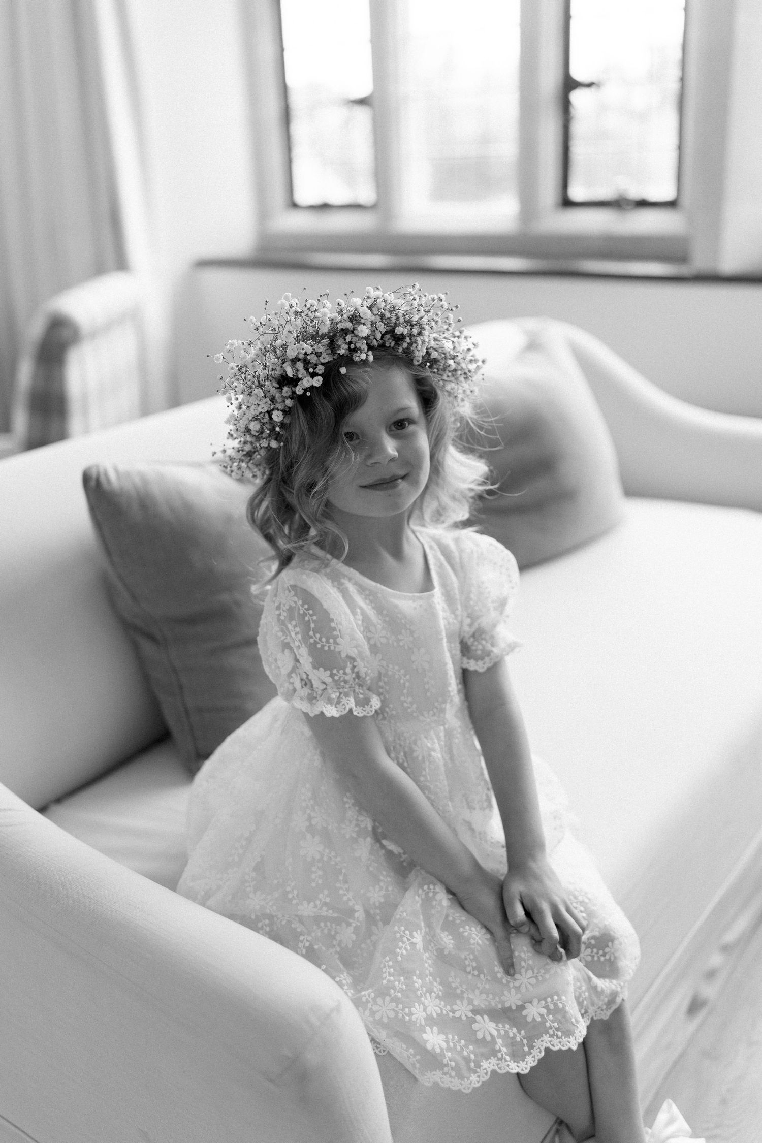 Black and white portrait of flower girl in white dress wearing daisy flower crown on sofa.