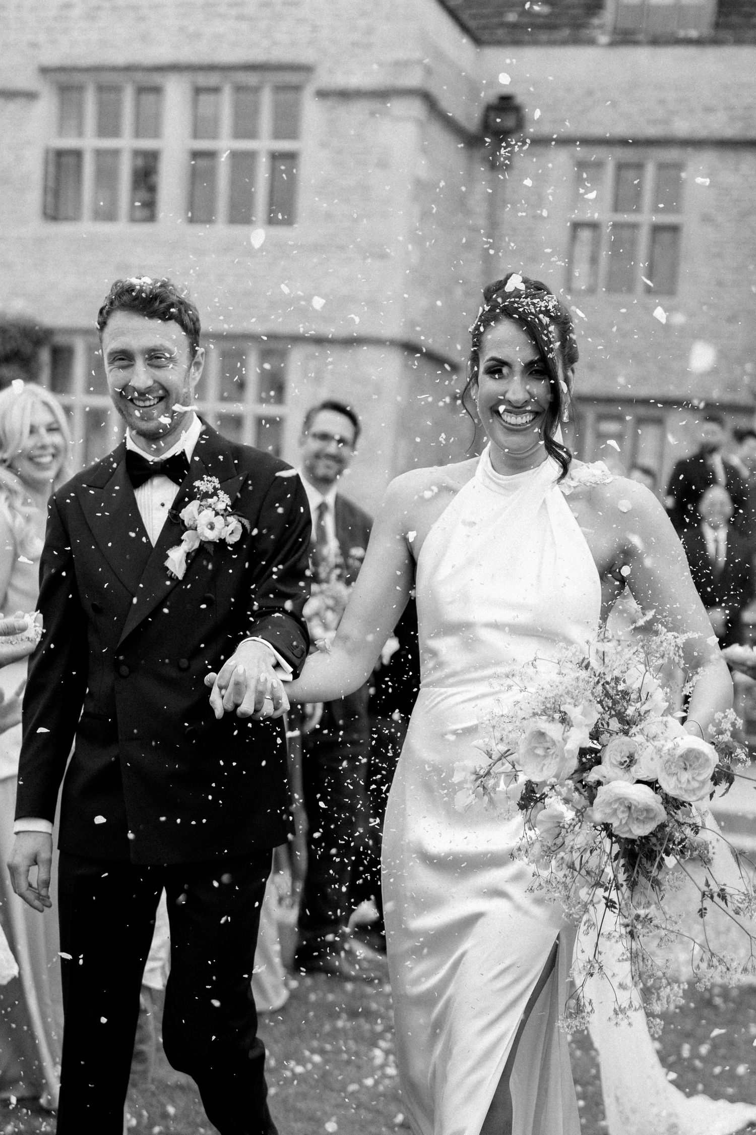 Joyful bride and groom laughing as confetti falls outside a historic stone manor house.