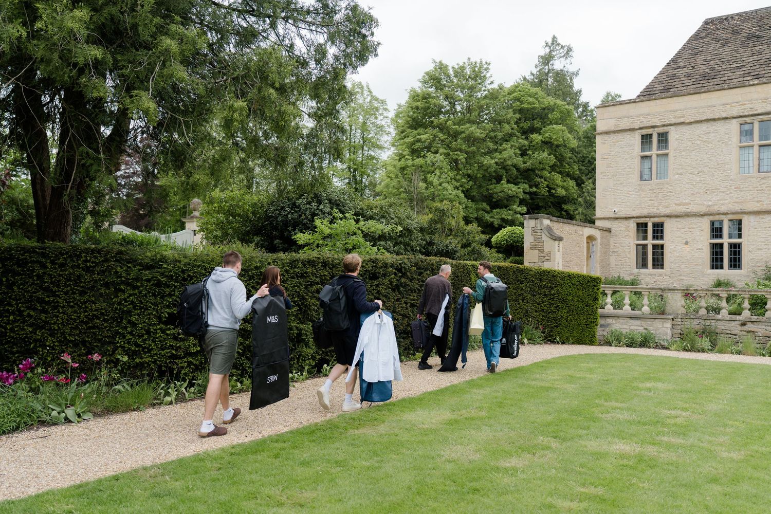 Wedding party guests walking along a garden path carrying garment bags at a stone manor house.