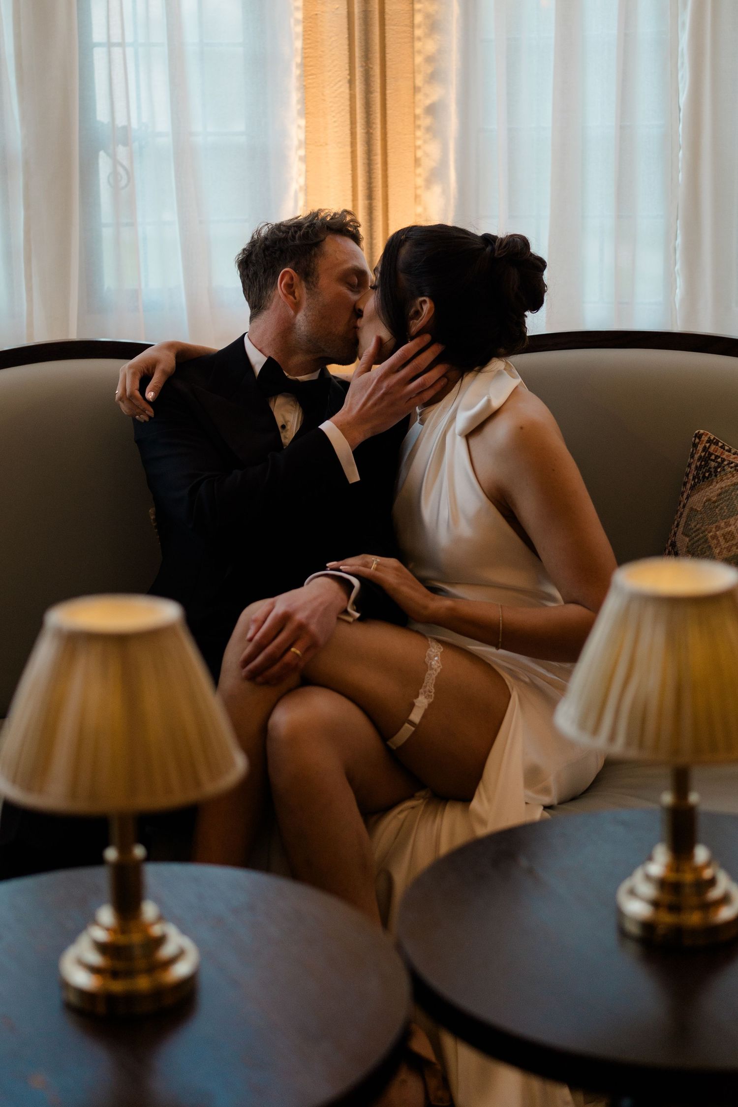 Bride and groom sharing romantic kiss on sofa in warmly lit room, bride wearing garter and short dress.