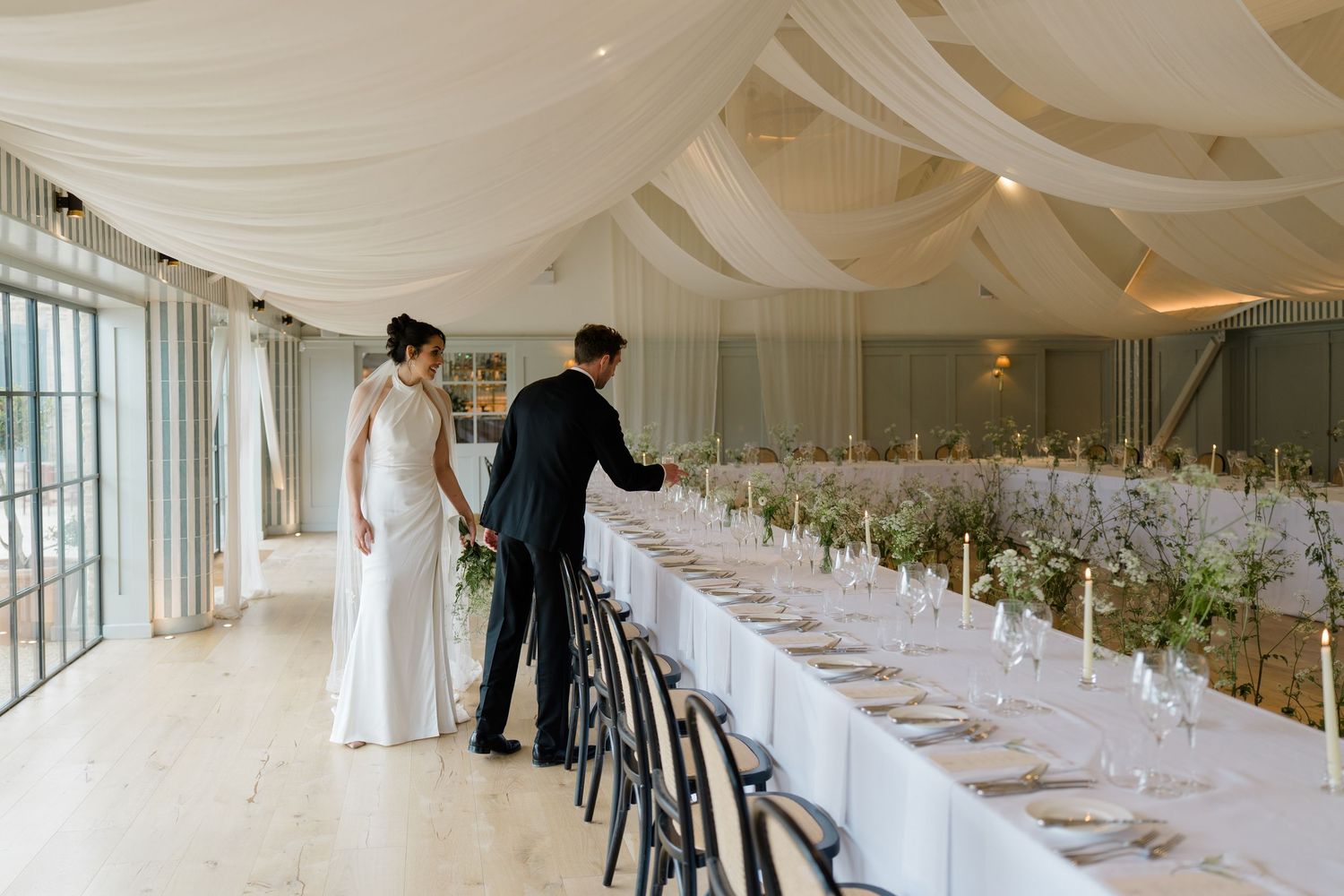 Bride and groom admire elegantly set long reception table beneath white draped ceiling in bright wedding venue.