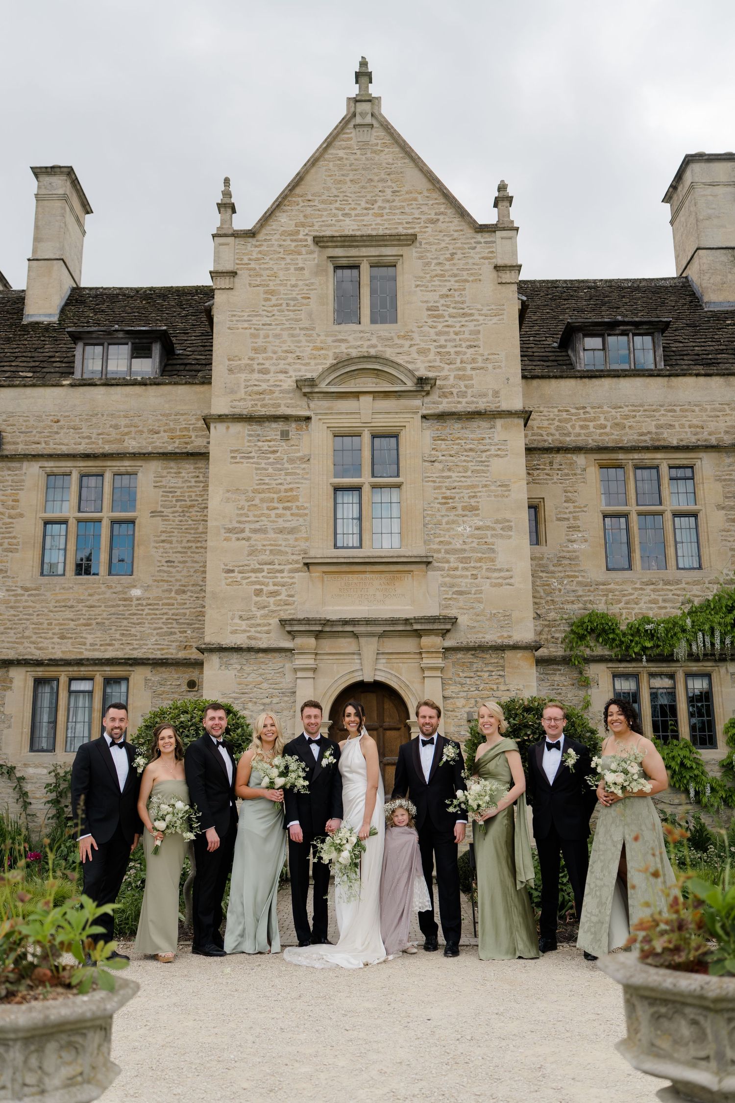 Wedding party poses before stone manor house, bridesmaids in sage green gowns, groomsmen in black tuxedos.