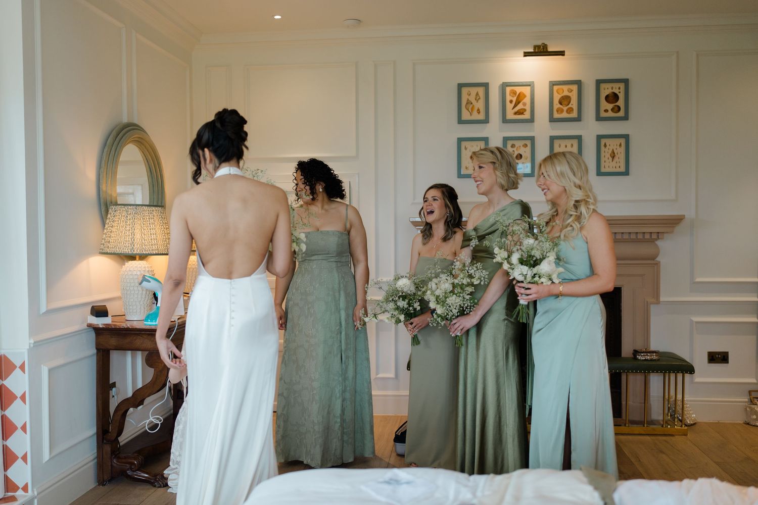 Bride in backless white gown stands with four bridesmaids in sage green dresses holding floral bouquets.