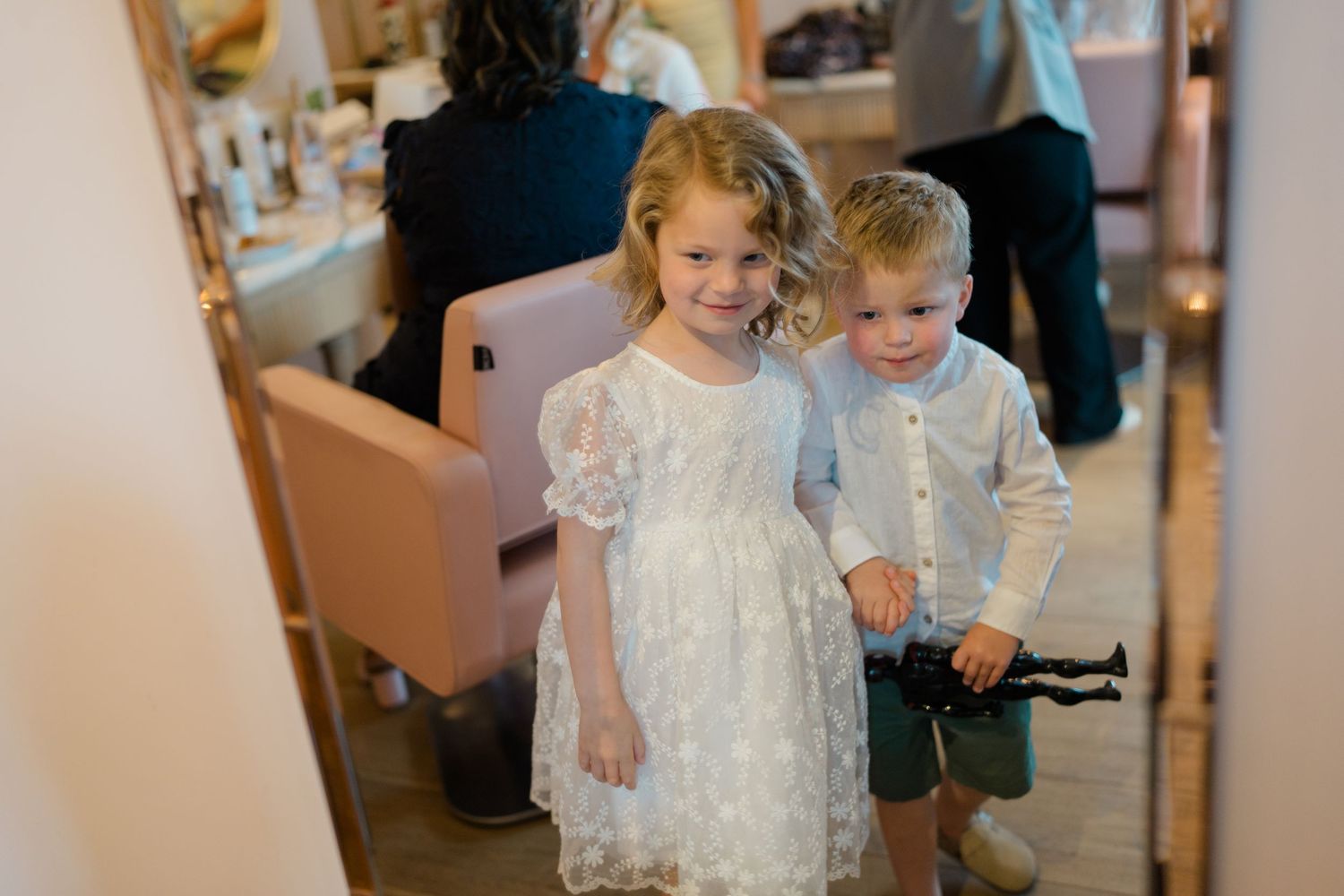 Two young children in formal white attire smile at a wedding getting ready venue.