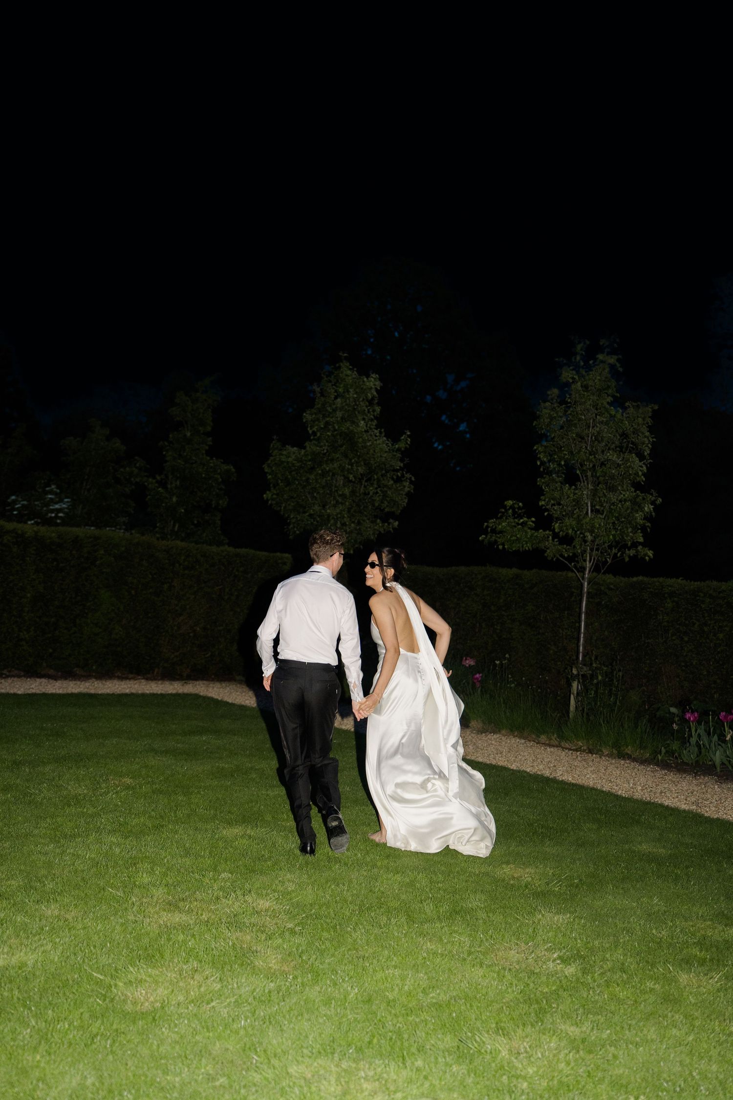 Bride and groom walking hand in hand across a moonlit garden at night after their wedding reception.