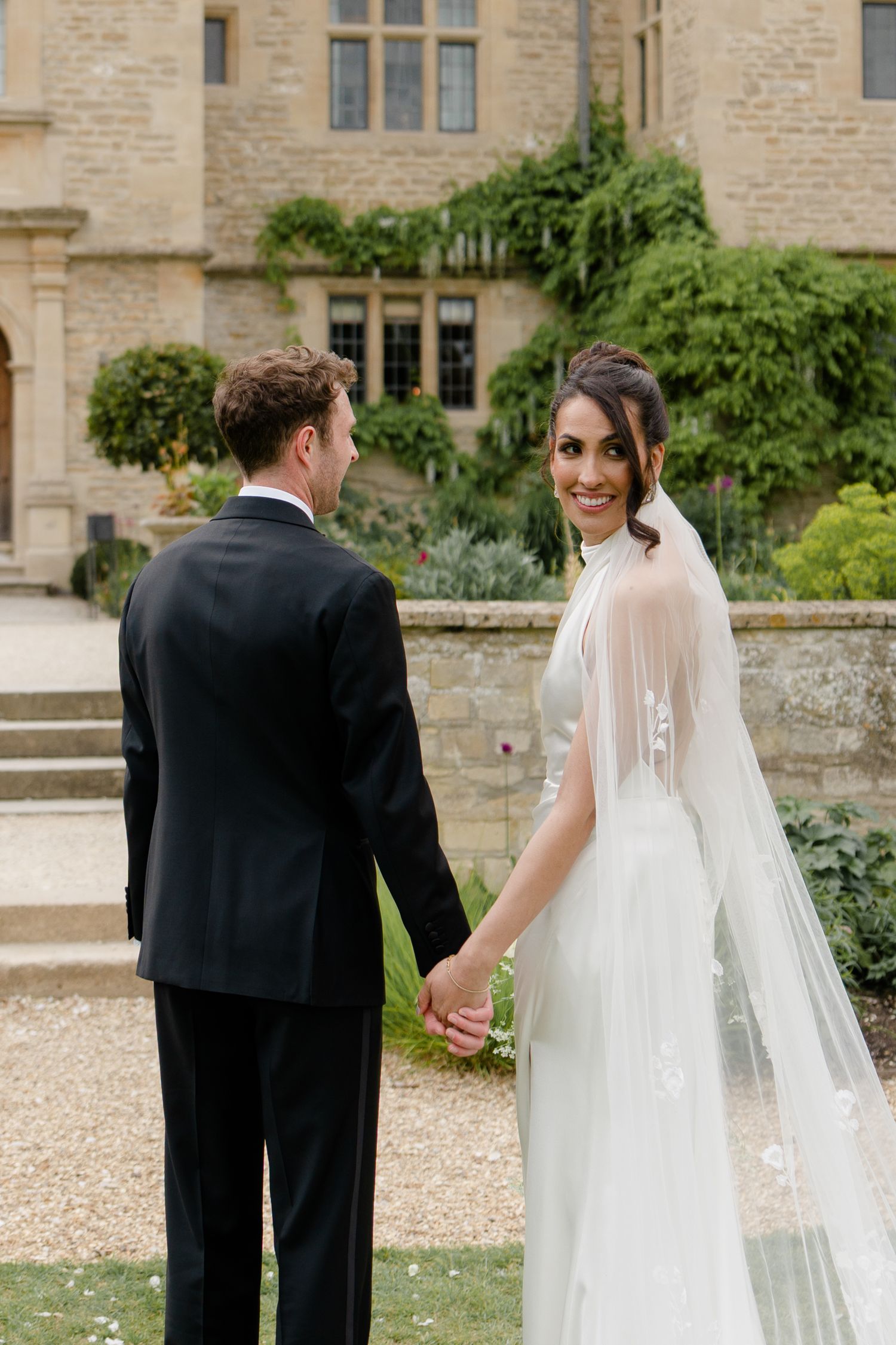 Smiling bride in white halter gown with veil holds grooms hand outside stone manor house.