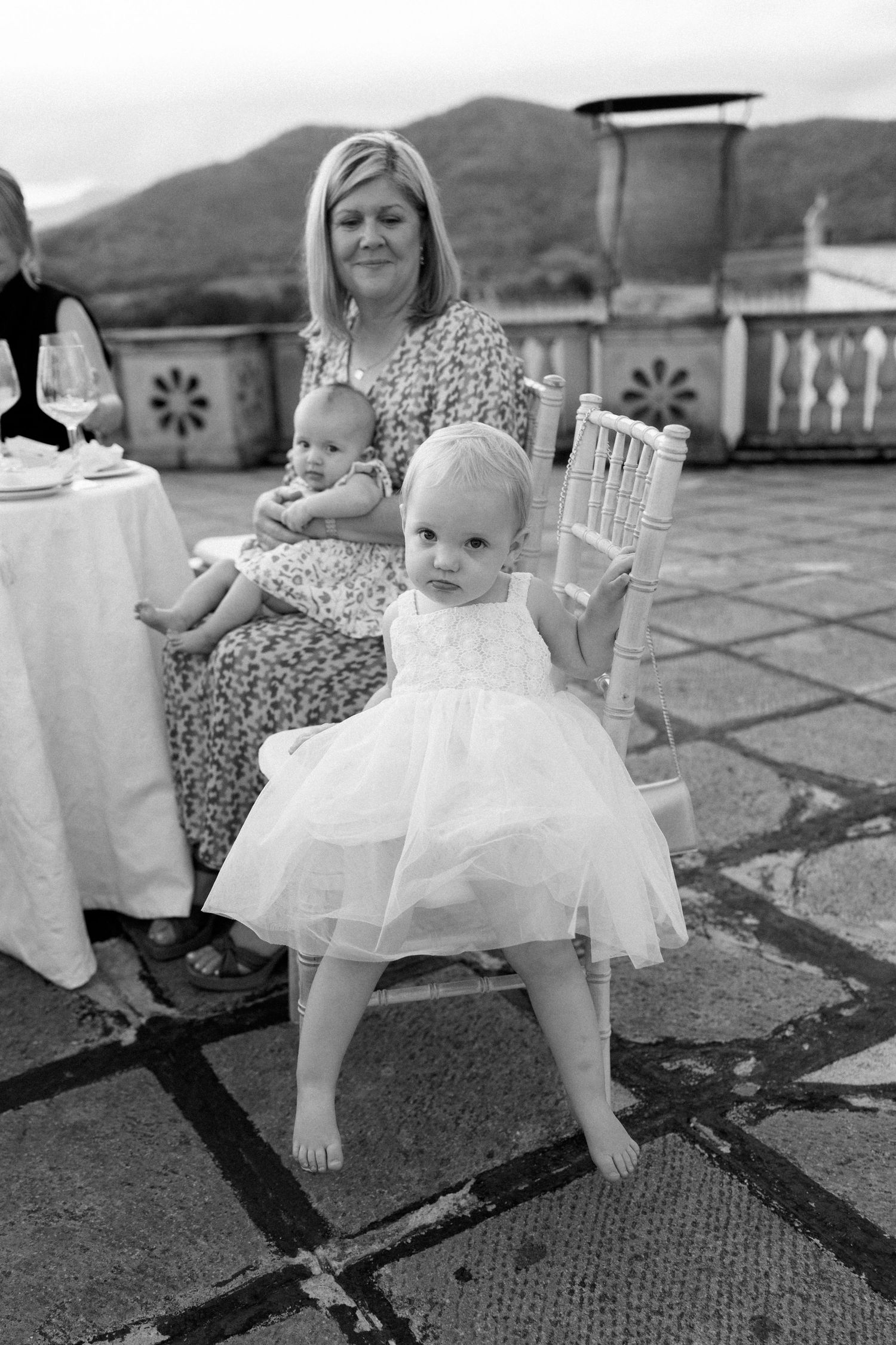 A toddler in a white tulle dress stands on a stone patio with mountain views during an outdoor gathering.