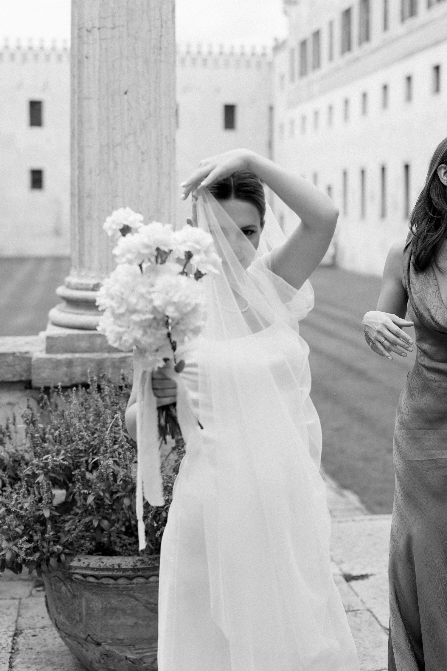 Black and white photo of bride with veil holding bouquet next to stone column.
