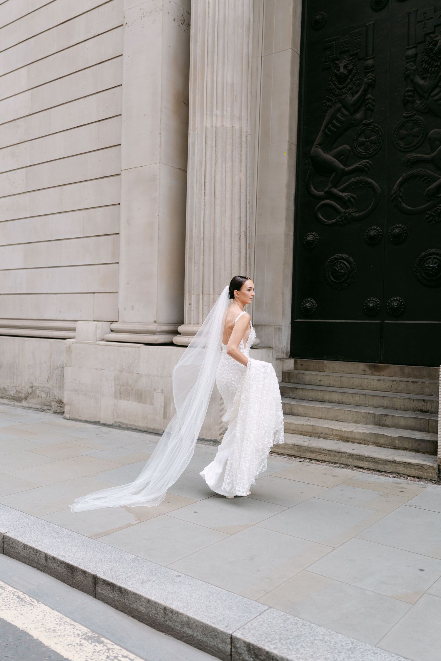 Bride walks gracefully along classical building facade with flowing veil.
