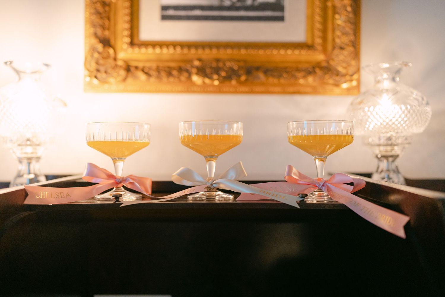 Four vintage coupe glasses with pink ribbons arranged in a row against a dark background.