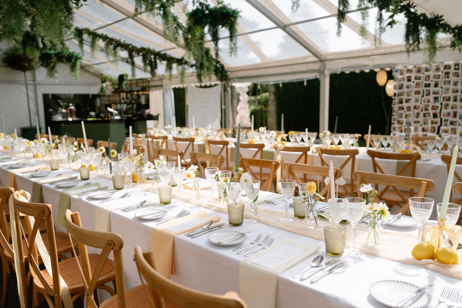 Wedding reception tables arranged under a clear tent with hanging greenery and warm lighting.
