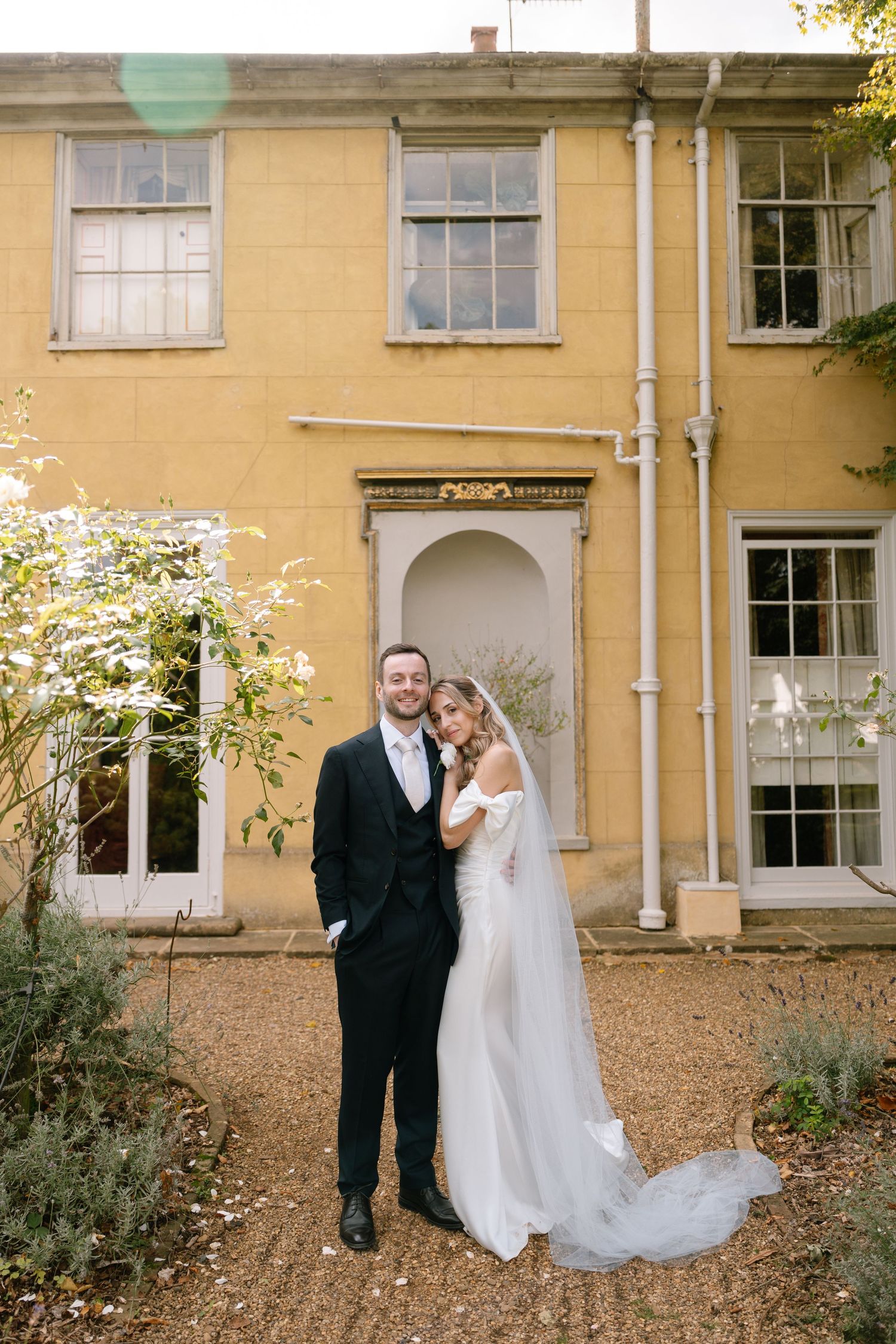 Wedding couple poses against yellow building facade with garden elements.