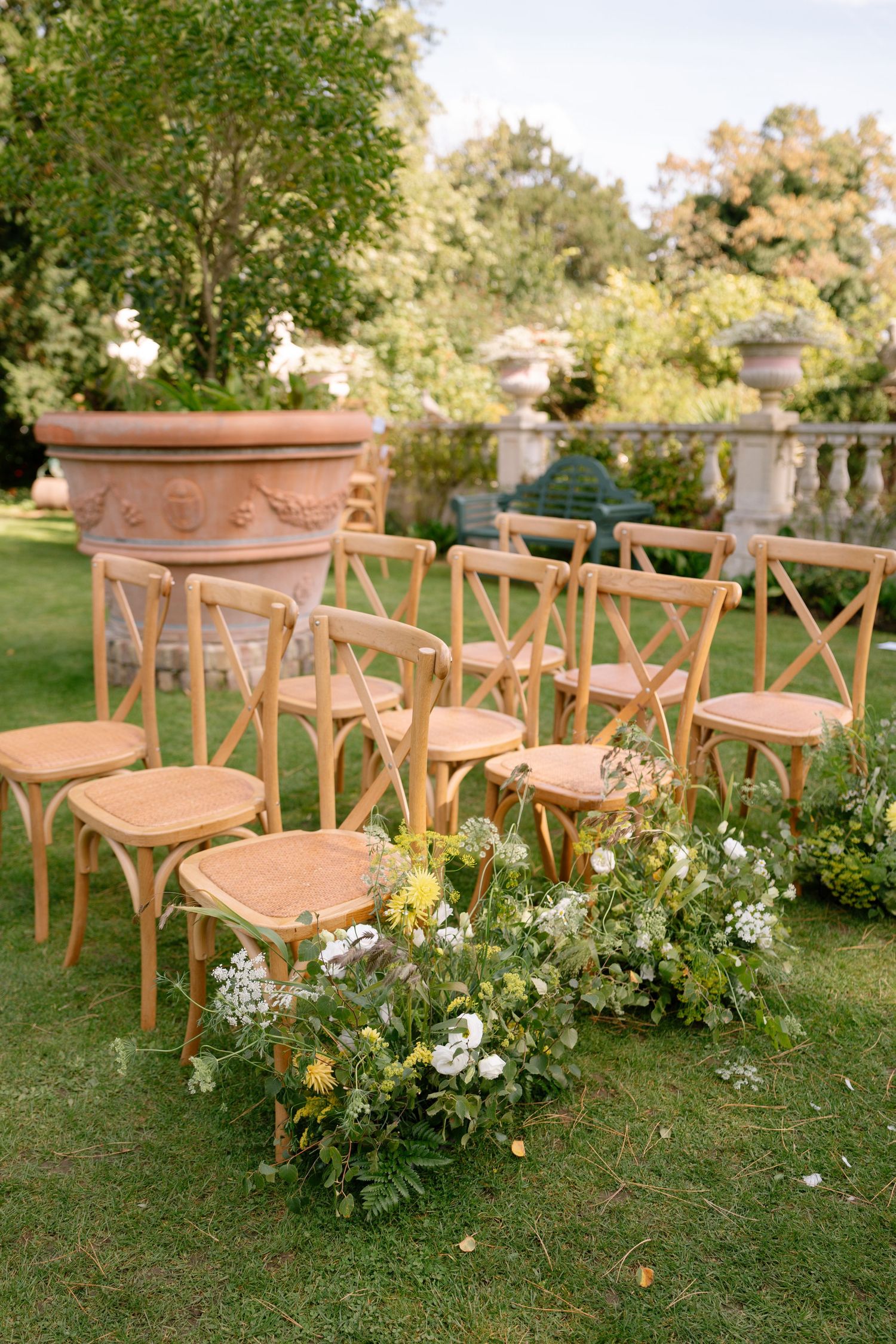 Wooden cross-back chairs arranged on a lawn with floral aisle decorations.