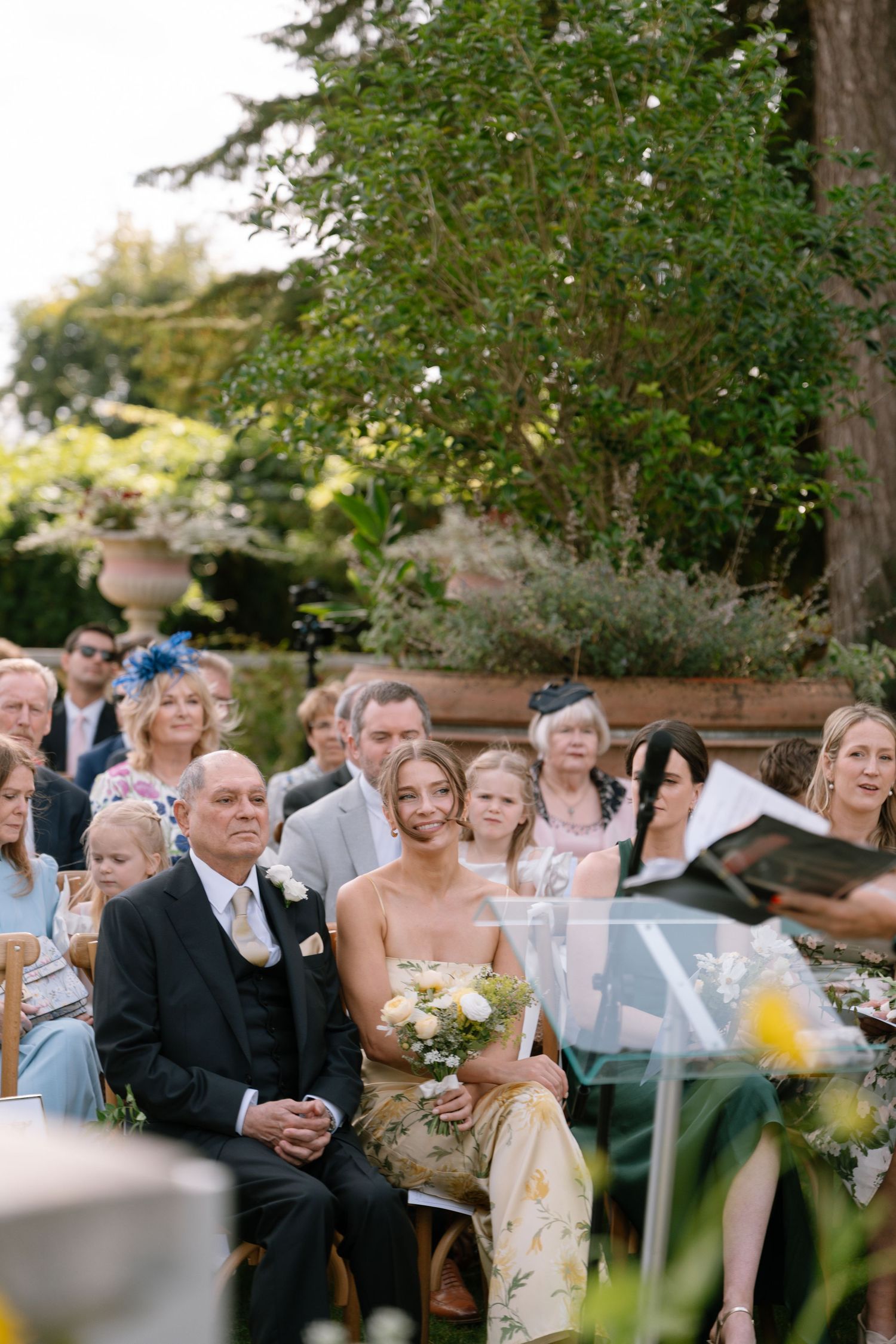 Wedding guests watch the ceremony from wooden chairs in a lush garden setting.