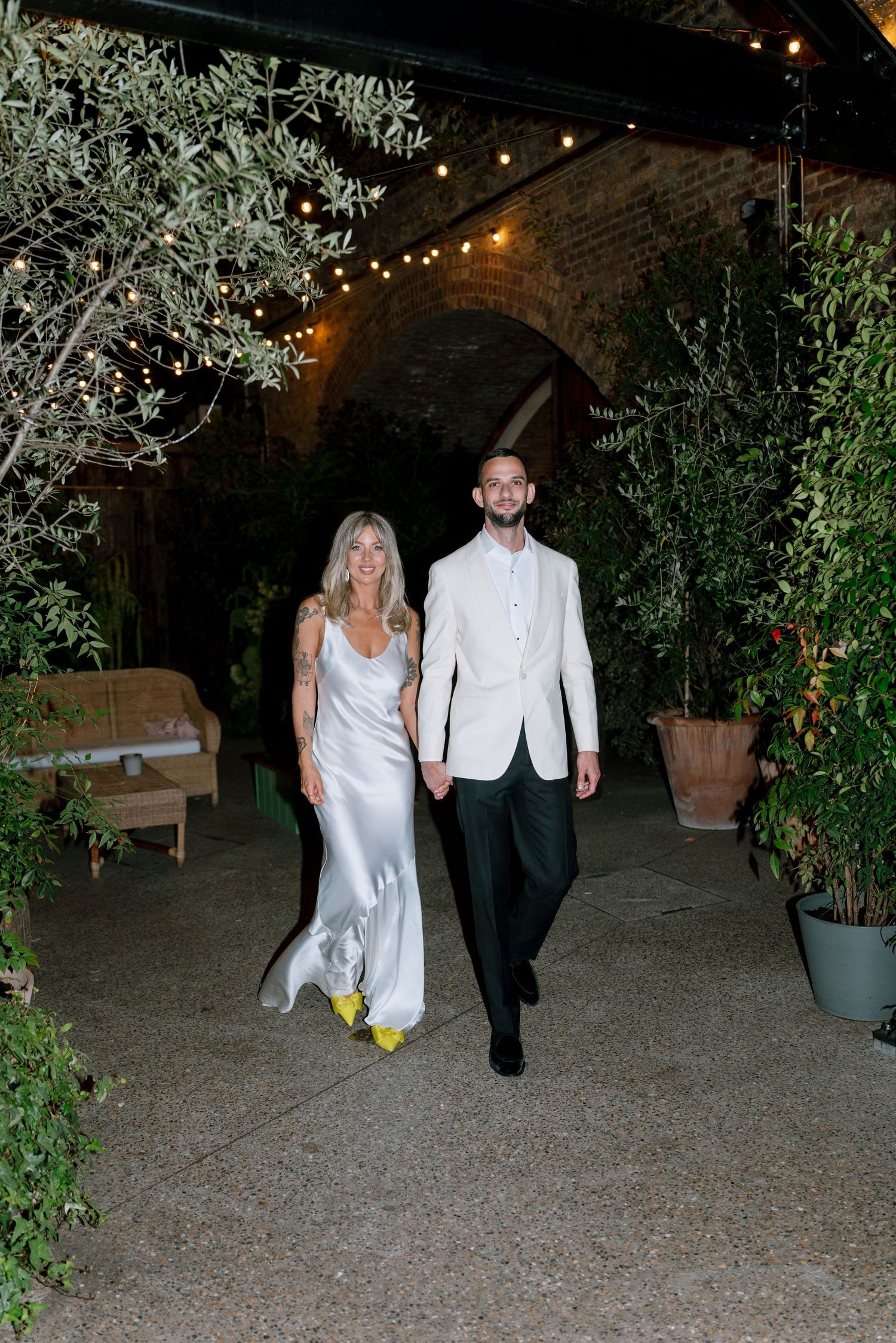 Couple in white formal attire walking together through an outdoor venue decorated with string lights.