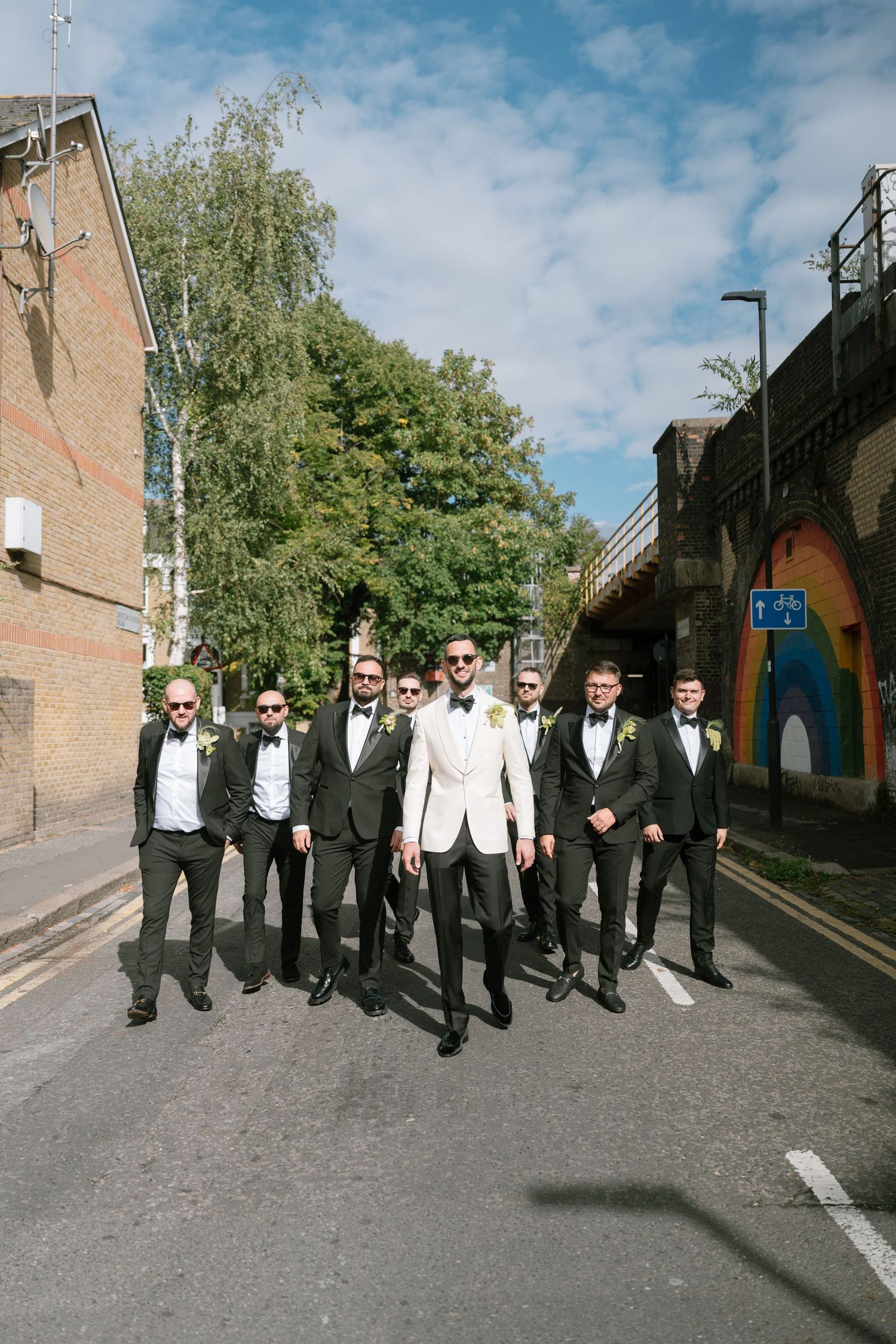 Groomsmen in formal attire walking down street with groom in white jacket.