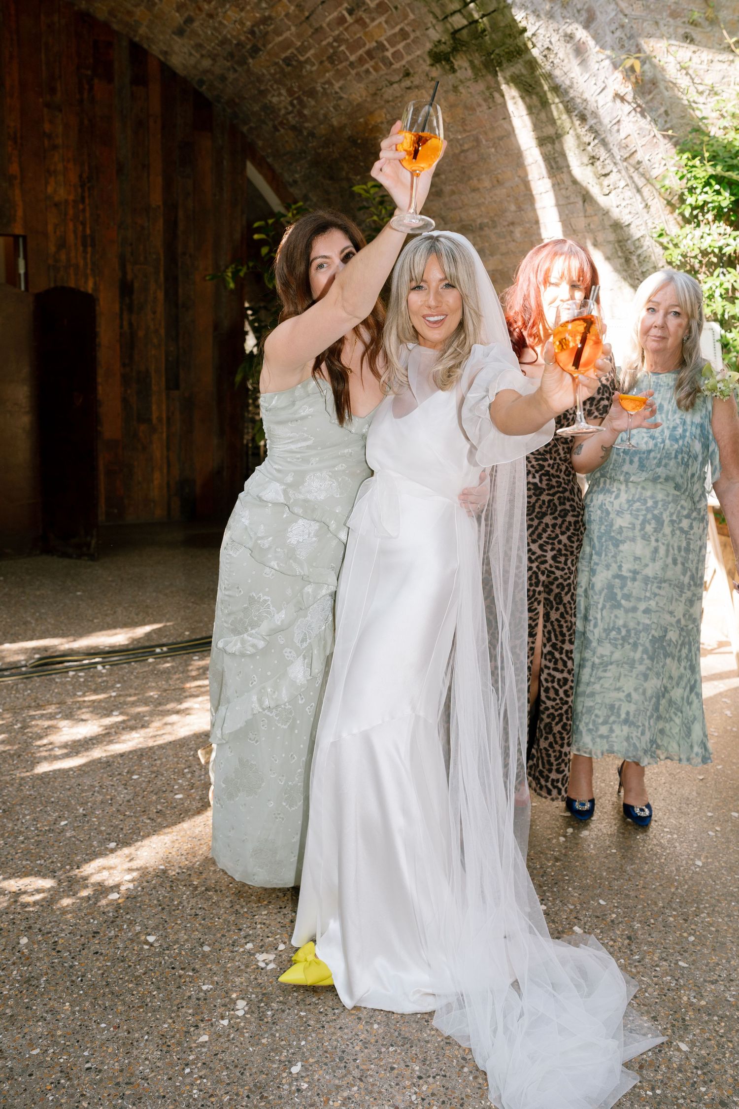 Wedding party raises champagne glasses in celebration under brick archway on sunny day.