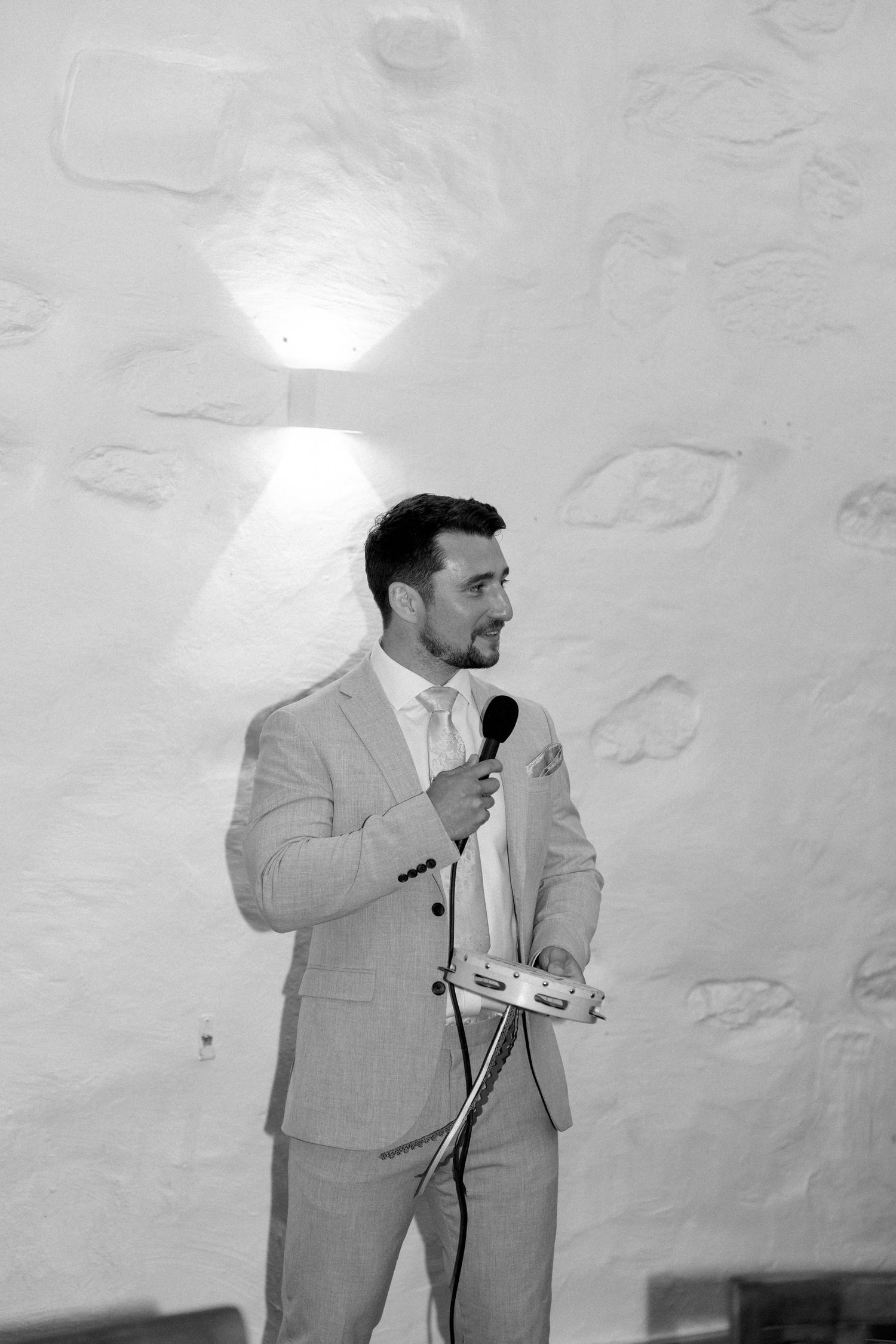 Man in light gray suit gives speech with microphone against white textured wall.