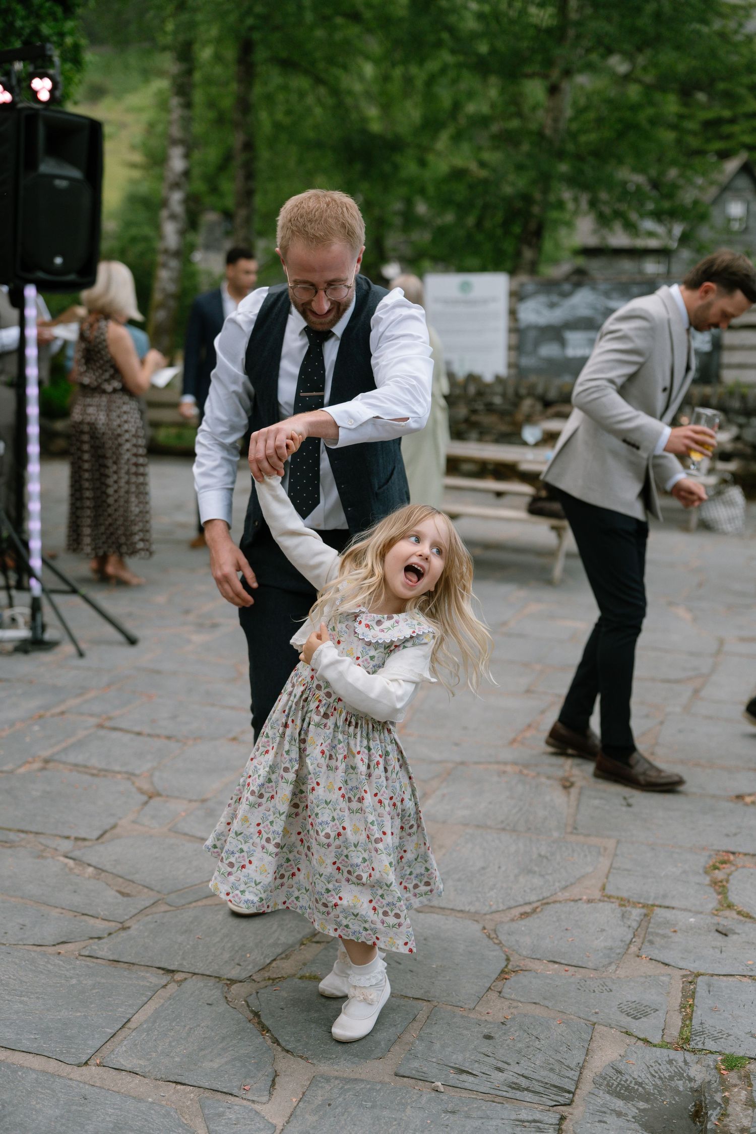 Young child in floral dress dances joyfully on stone patio during wedding reception.