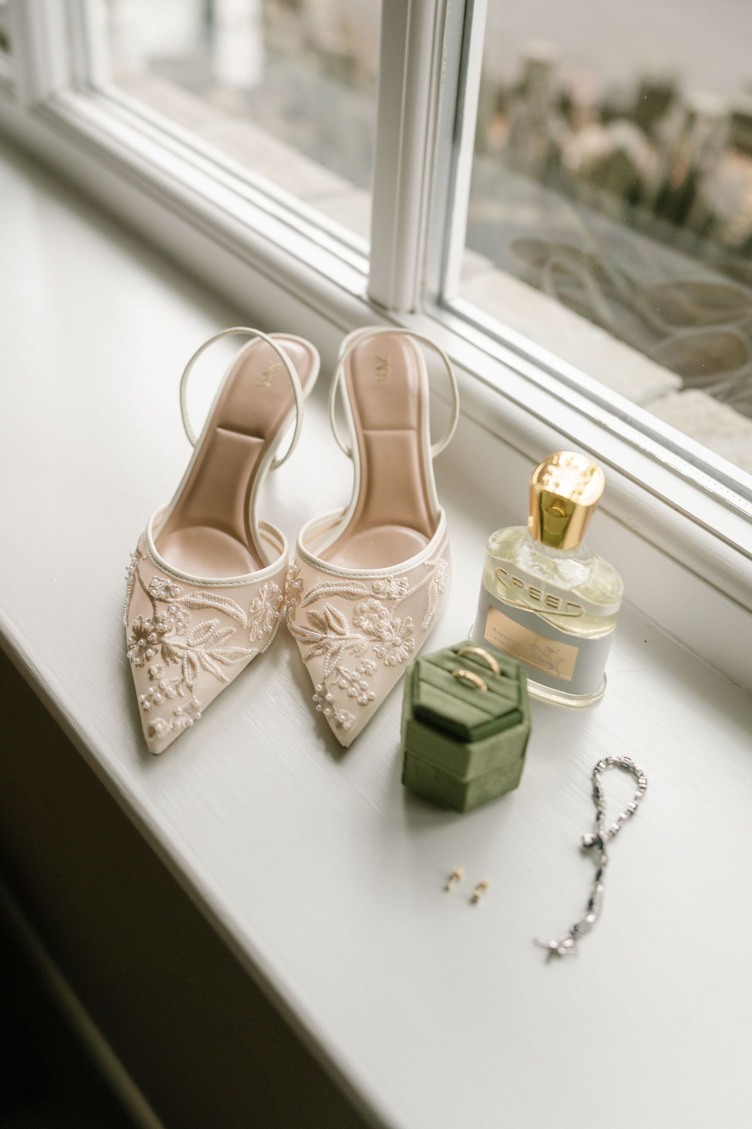Delicate lace bridal shoes displayed on a white windowsill next to a dark green velvet ring box.