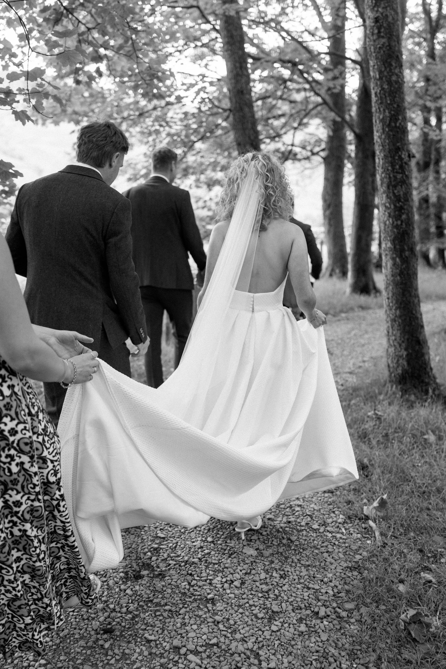 Wedding party walks through a wooded area with flowing white dress fabric trailing behind.