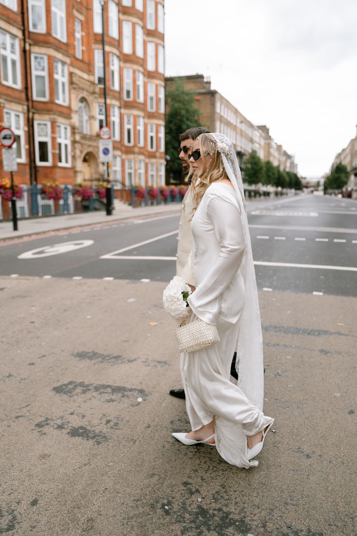 A bride in a modern white pantsuit and veil poses on a London street with classic brick buildings.