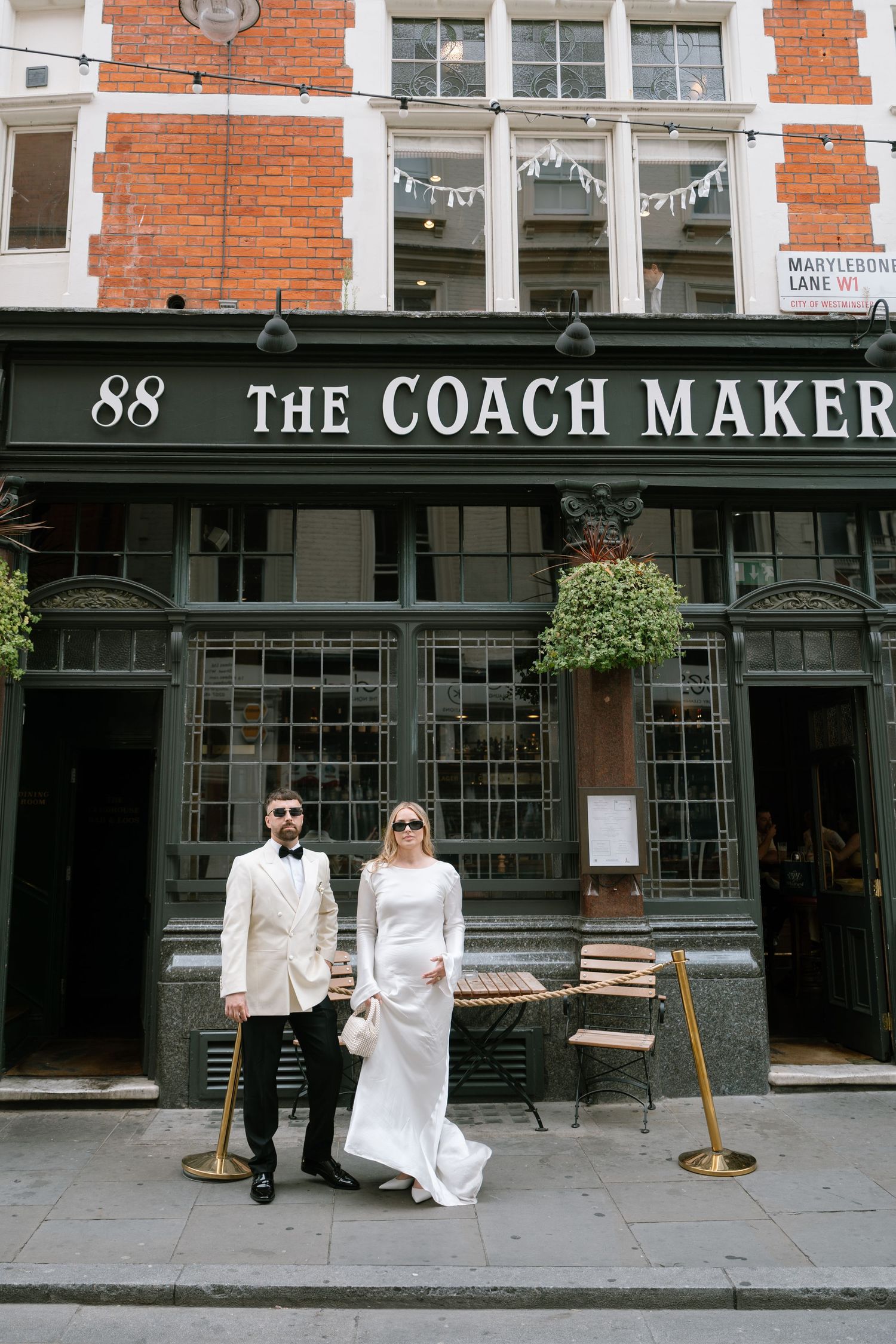 A couple poses in formal white attire outside The Coach Makers pub with classic brick architecture.